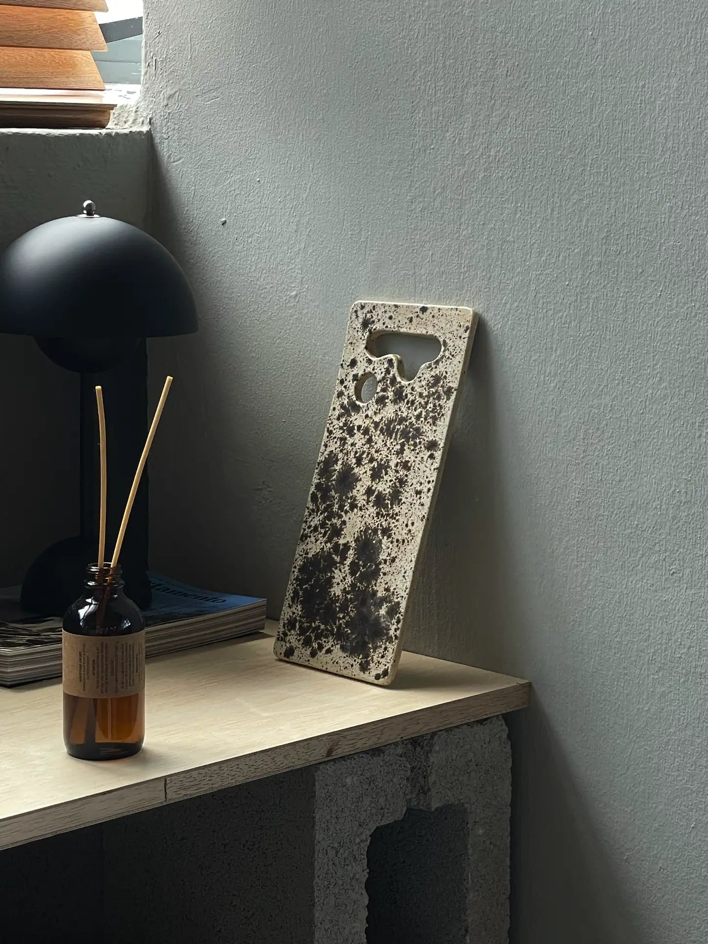 A speckled phone case rests on a wooden desk near a reed diffuser, black lamp, and magazines, evoking the rustic texture and unique cutouts of the Mirjam Faber Ceramics Sushi Plate by Mirjam Faber Ceramics.