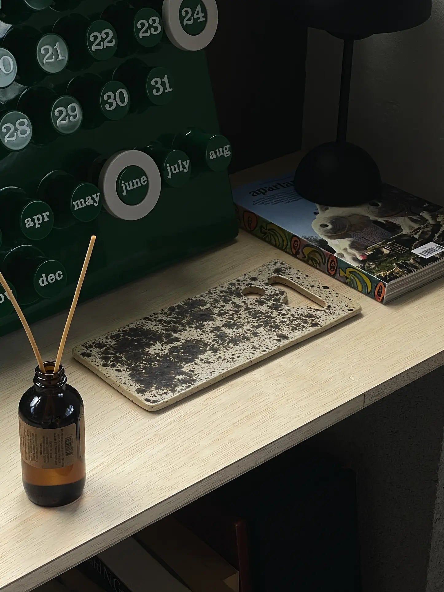 A wooden desk holds a cutting board, a brown reed diffuser, the Mirjam Faber Ceramics Sushi Plate by Mirjam Faber Ceramics with unique cutouts, a green perpetual calendar, and an animal-themed magazine. Sunlight adds calm to the scene.