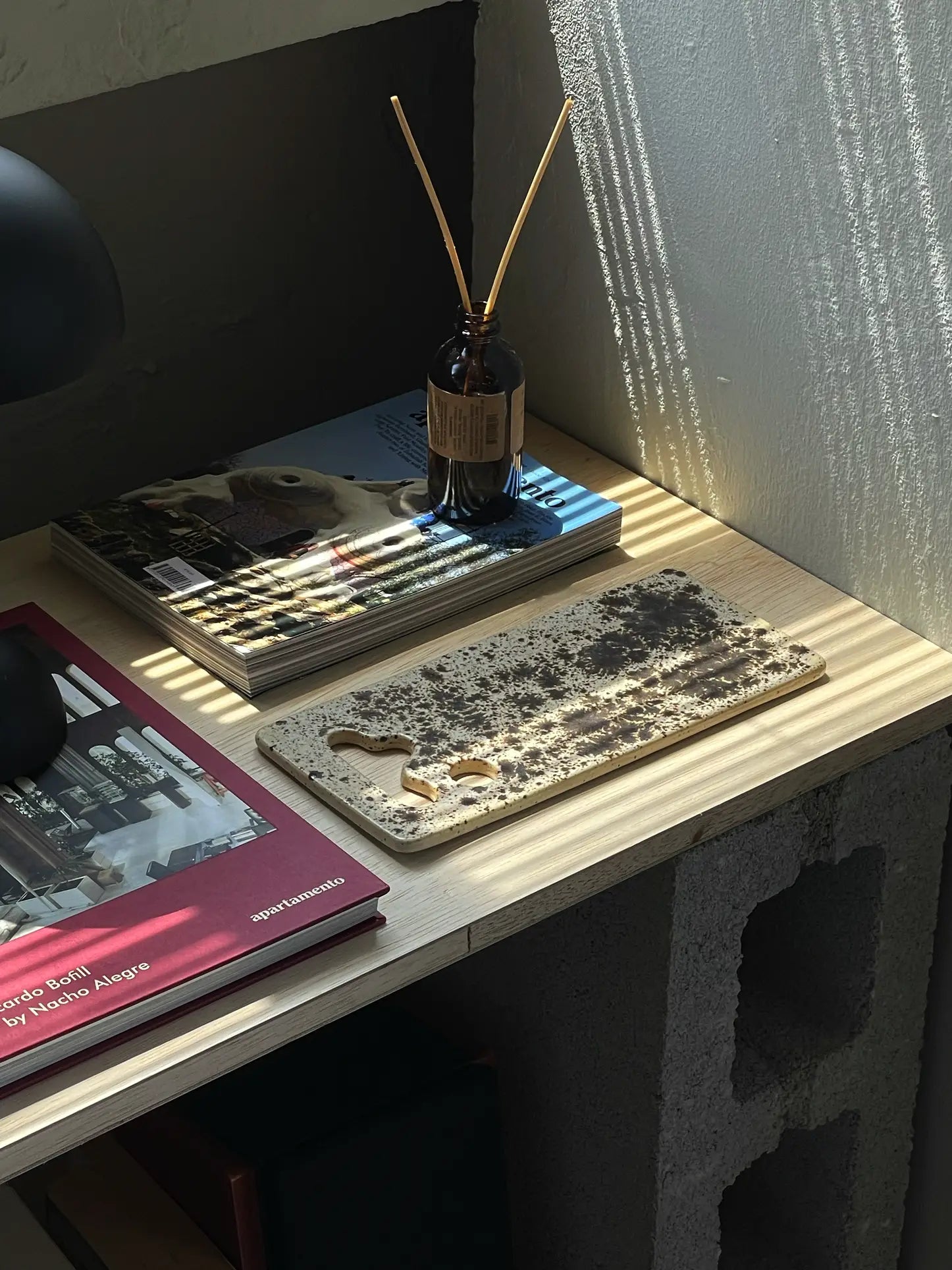 A wooden desk with sunlight streaming in displays a red book, a magazine, a Mirjam Faber Ceramics Sushi Plate by Mirjam Faber Ceramics, a speckled cutting board, and a brown glass bottle with reed diffusers on a concrete block base.