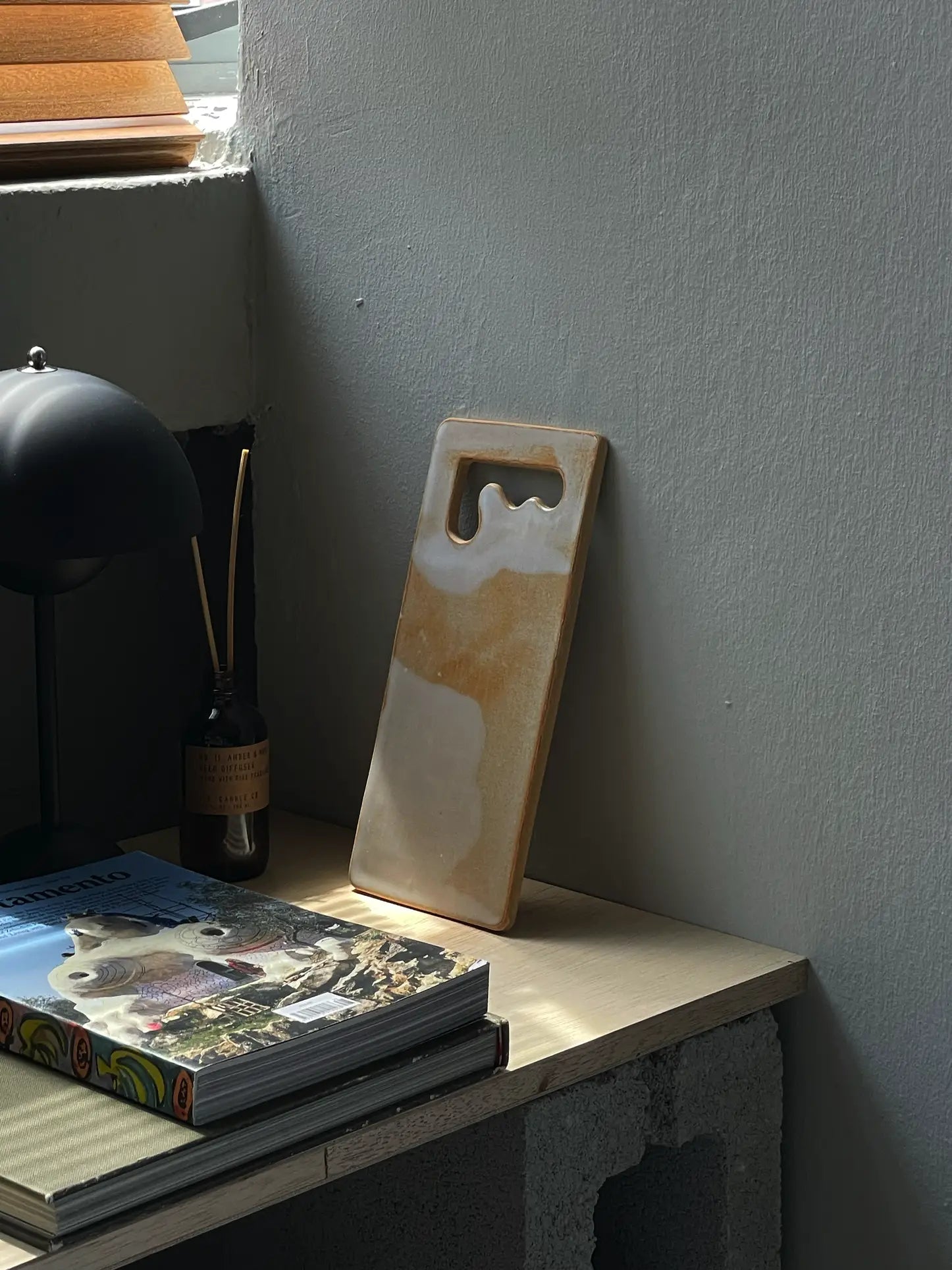 The Mirjam Faber Ceramics Sushi Plate, featuring abstract beige and white patterns, leans against a gray wall on a wooden desk by a window, beside a book, lamp, and reed diffuser.