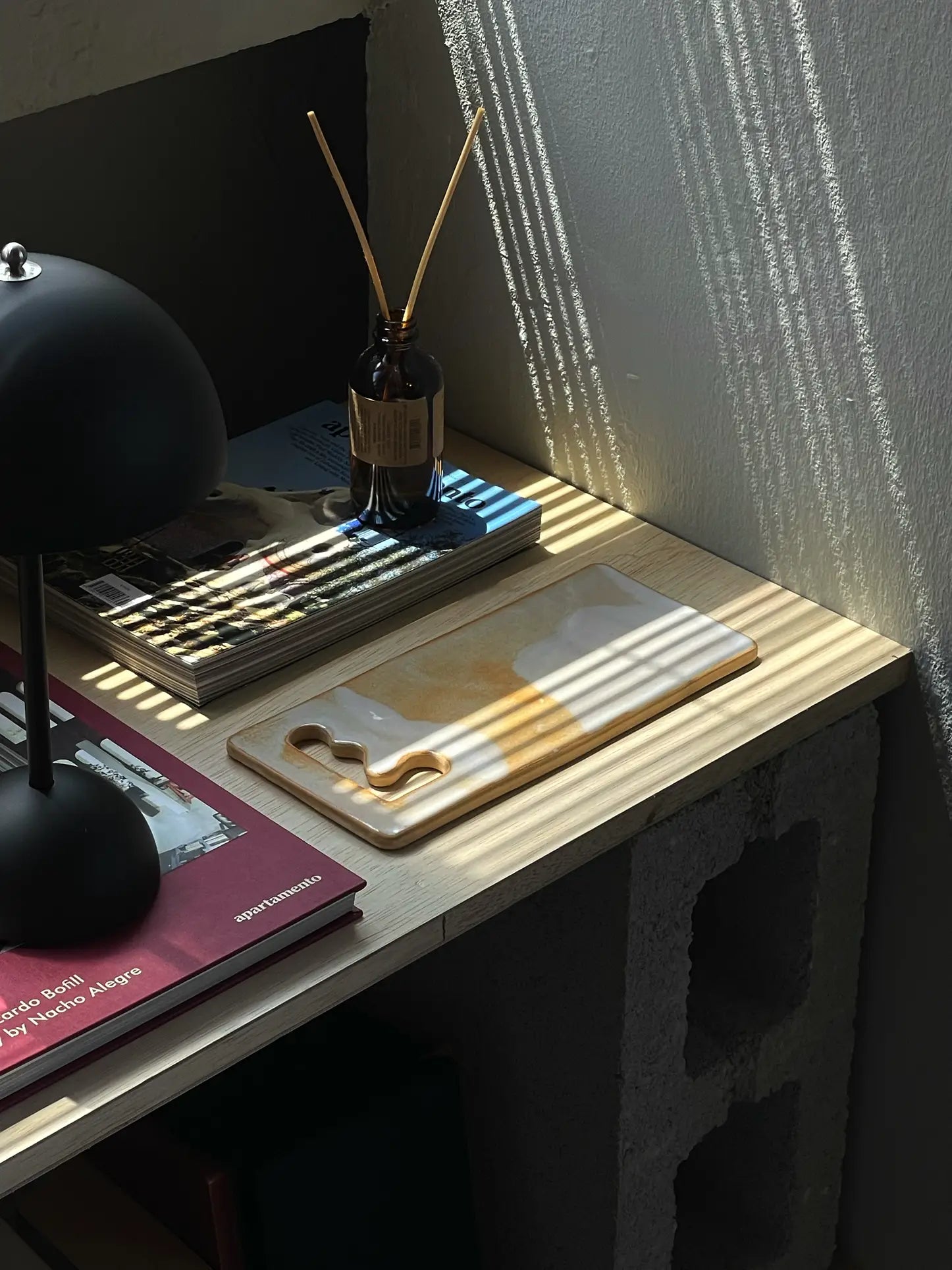 A sunlit desk with a black lamp, brown glass diffuser, magazines, and a Mirjam Faber Ceramics Sushi Plate featuring unique cutouts. Sunlight casts striped shadows through blinds onto the light wood surface.