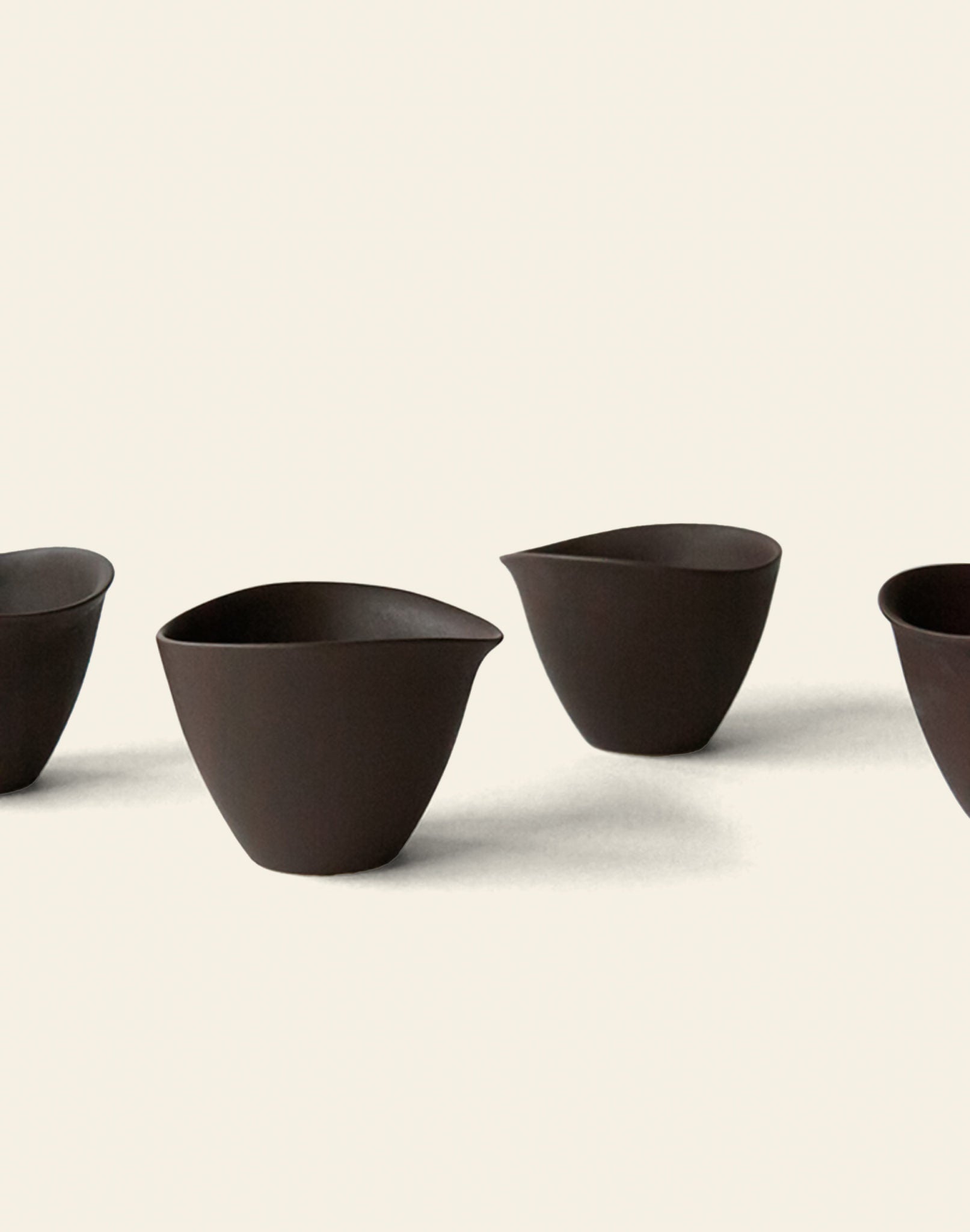 Four dark brown NR Ceramics HIN Tea Cups with a minimalist, ergonomic design and pointed lips are arranged on a light background, casting soft shadows for a clean, modern look.