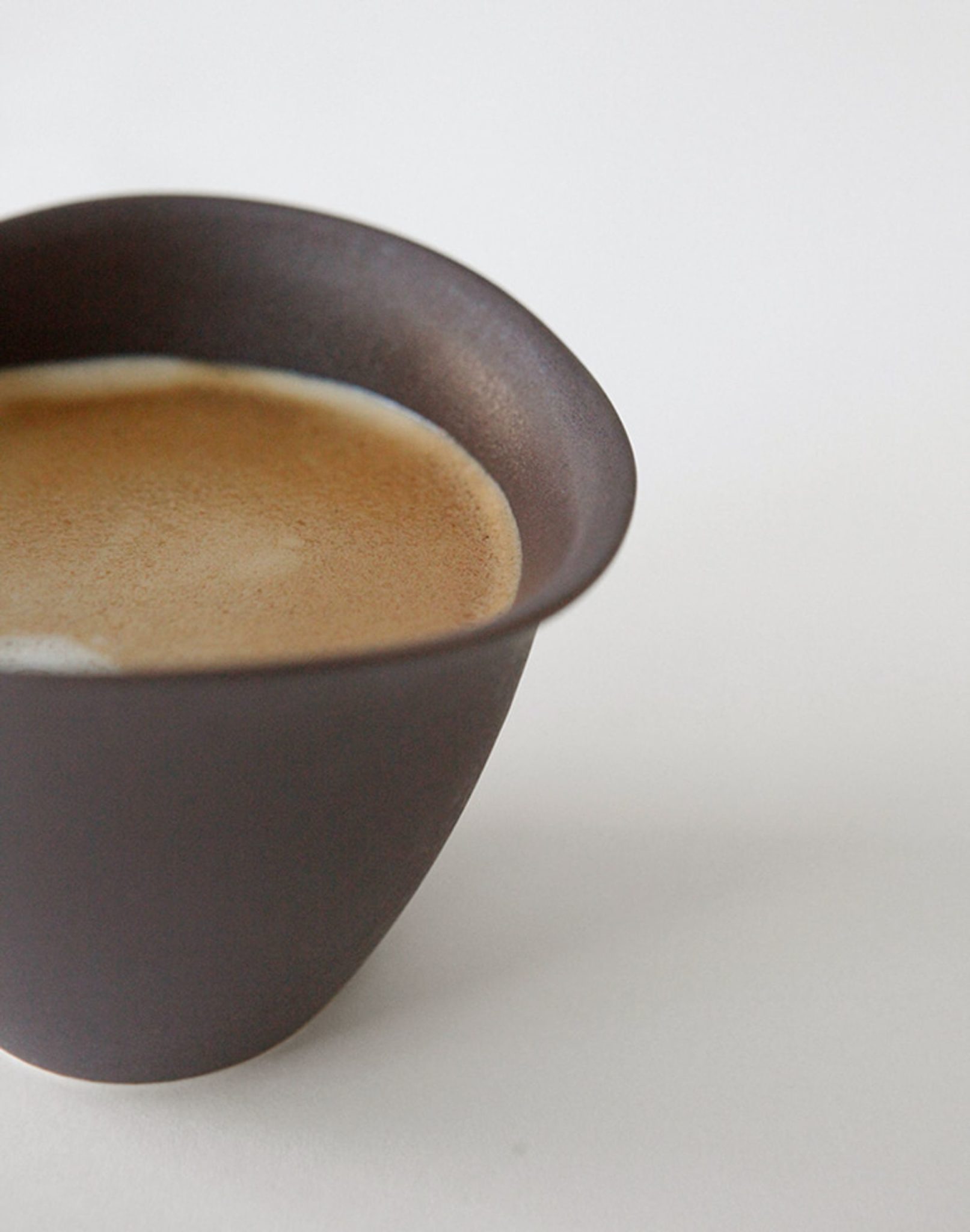 A close-up of the NR Ceramics HIN Tea Cup by NR Ceramics, a dark matte ceramic cup with an ergonomic design, filled with coffee topped with foamy crema, set against a plain white background and partially out of frame on the left.