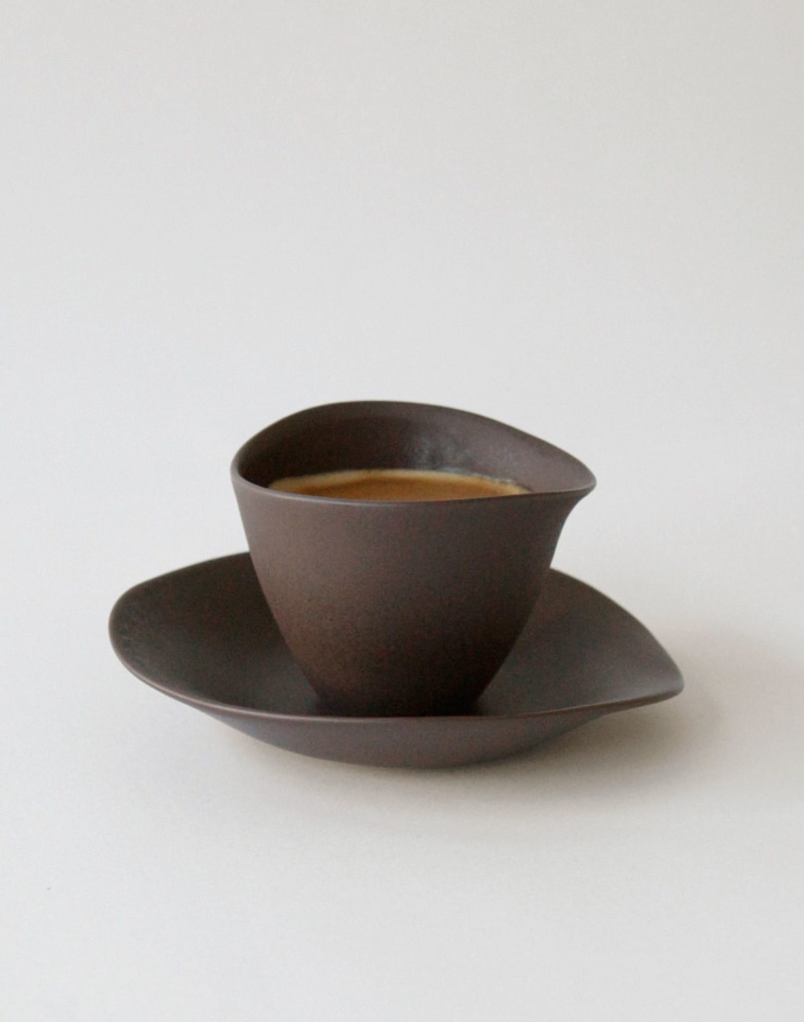 A modern, dark brown NR Ceramics HIN Tea Cup by NR Ceramics, filled with coffee and resting on a matching saucer, features an irregular asymmetrical shape and ergonomic design, both displayed against a plain white background.