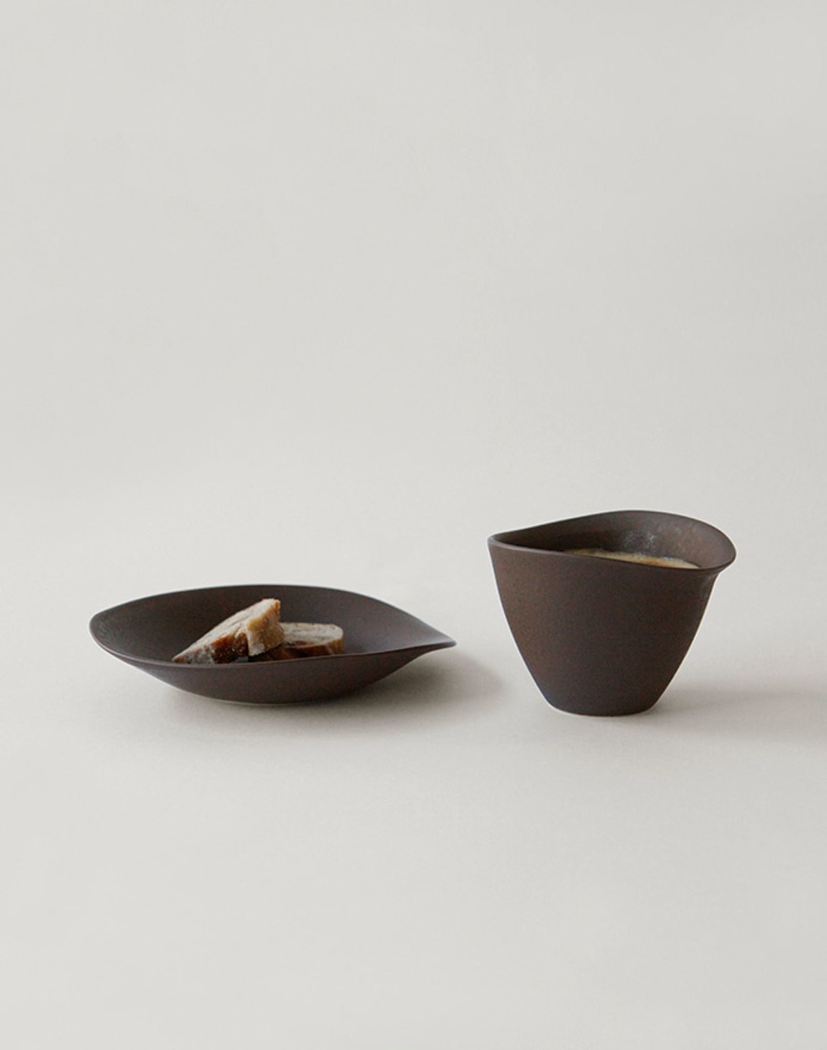 A dark minimalist plate holds three small breads next to the NR Ceramics HIN Tea Cup, both designed with ergonomic style by NR Ceramics, set on a plain light background.