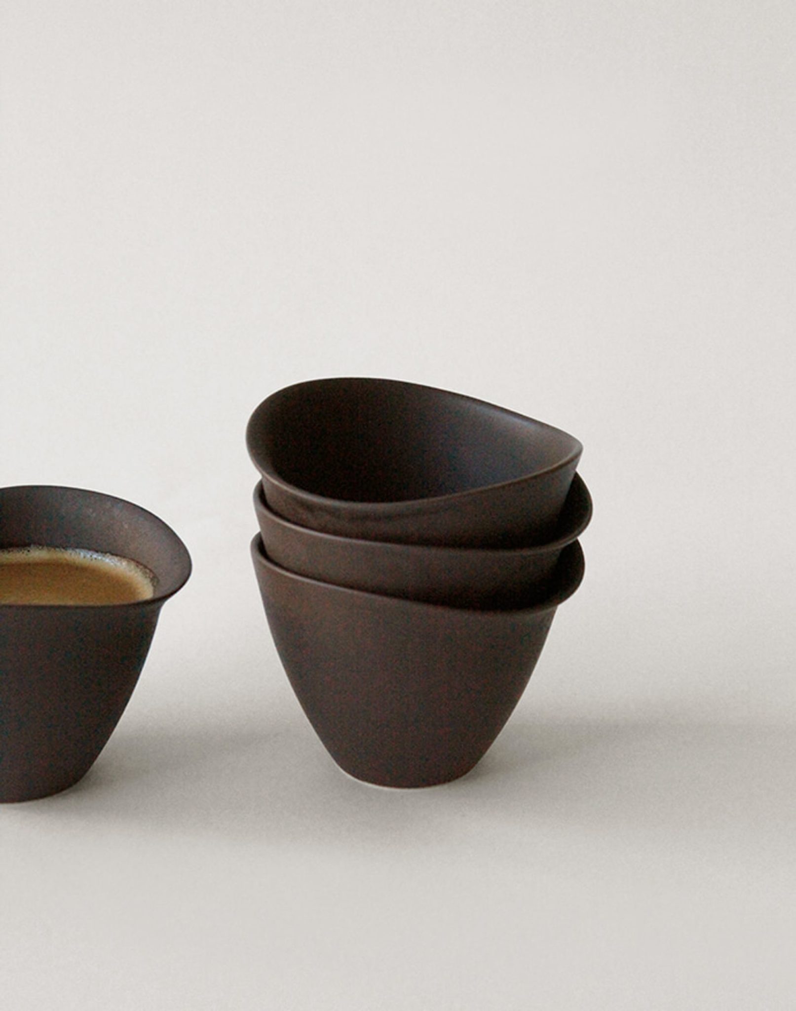 Three stacked NR Ceramics HIN Tea Cups sit beside another NR Ceramics HIN Tea Cup filled with coffee, all against a plain light background that highlights their ergonomic design.