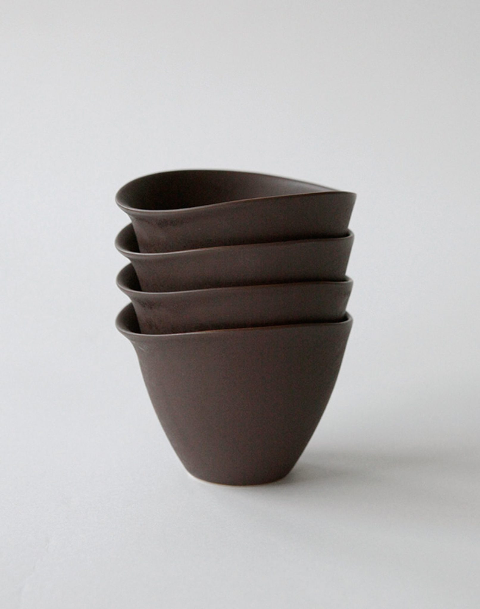 Four NR Ceramics HIN Tea Cups in dark brown, each featuring a minimalist and ergonomic design, are stacked neatly together by NR Ceramics against a plain, light gray background.