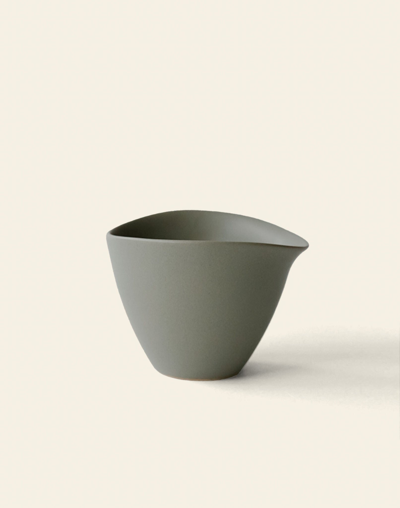 A matte gray NR Ceramics HIN Tea Cup with an ergonomic, asymmetrical design and a slightly pointed lip sits on a light, neutral background.