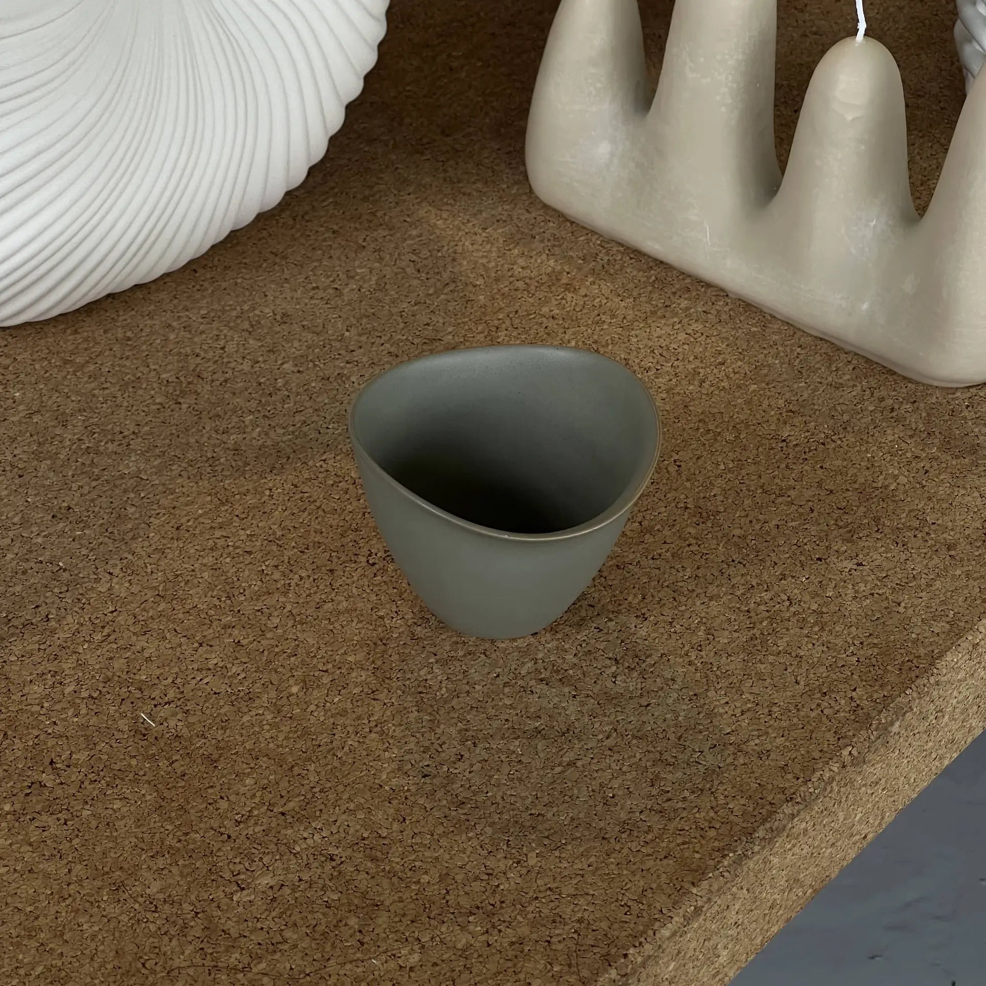A compact, matte gray NR Ceramics HIN Tea Cup with an ergonomic design rests on a cork surface next to a ribbed white vase and an abstract beige candle featuring four arches.