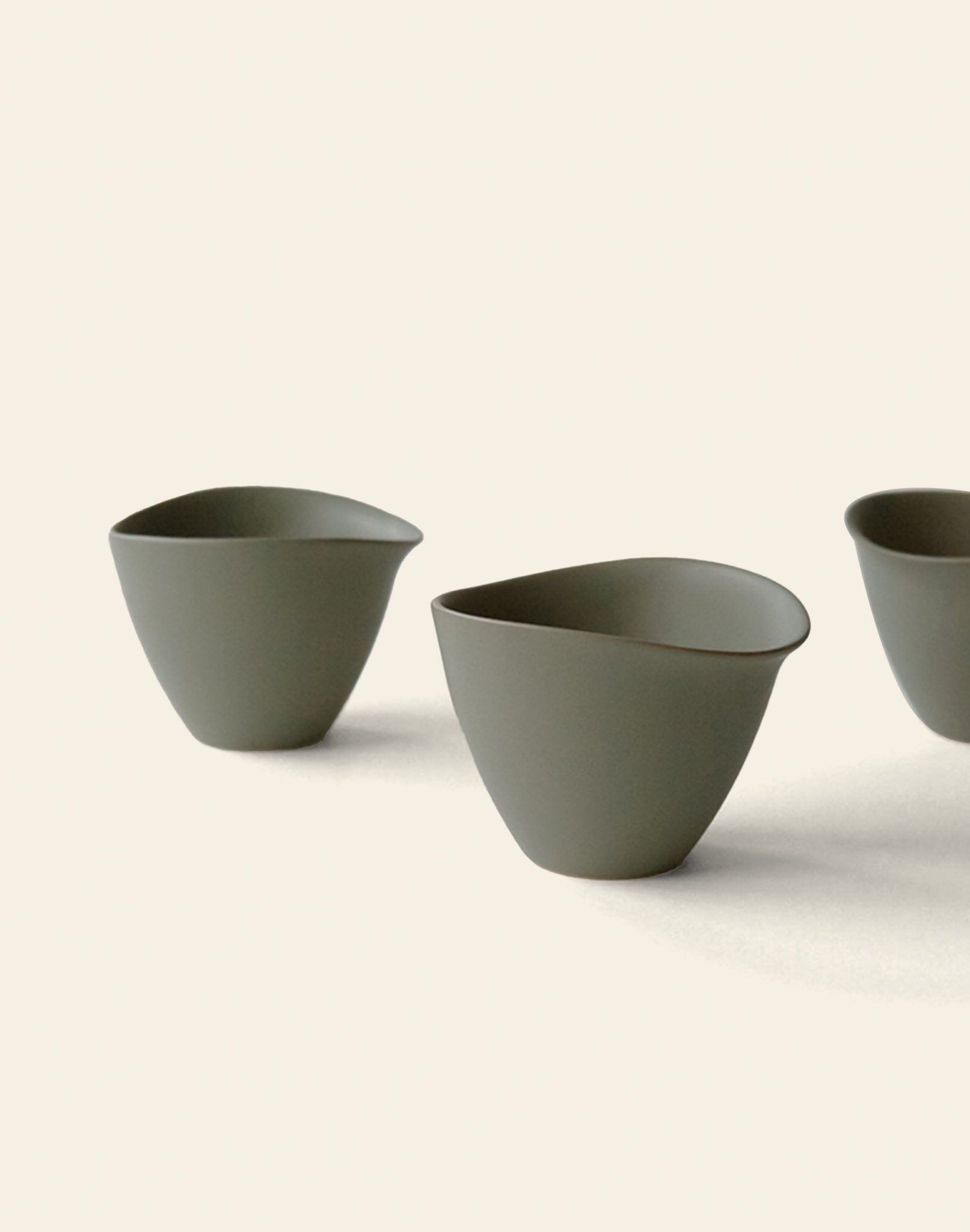 Three minimalist, matte olive green NR Ceramics HIN Tea Cups with ergonomic, organic shapes are arranged on a plain off-white background. Only part of the third cup on the right is visible.