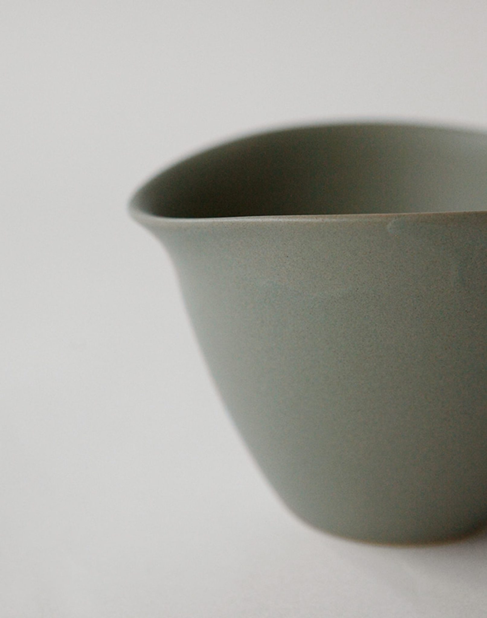 A close-up of the NR Ceramics HIN Tea Cup by NR Ceramics, featuring a simple matte grey finish, smooth surface, and slightly curved rim. The ergonomic design is highlighted against a light background, with the cup partially cropped on the right.