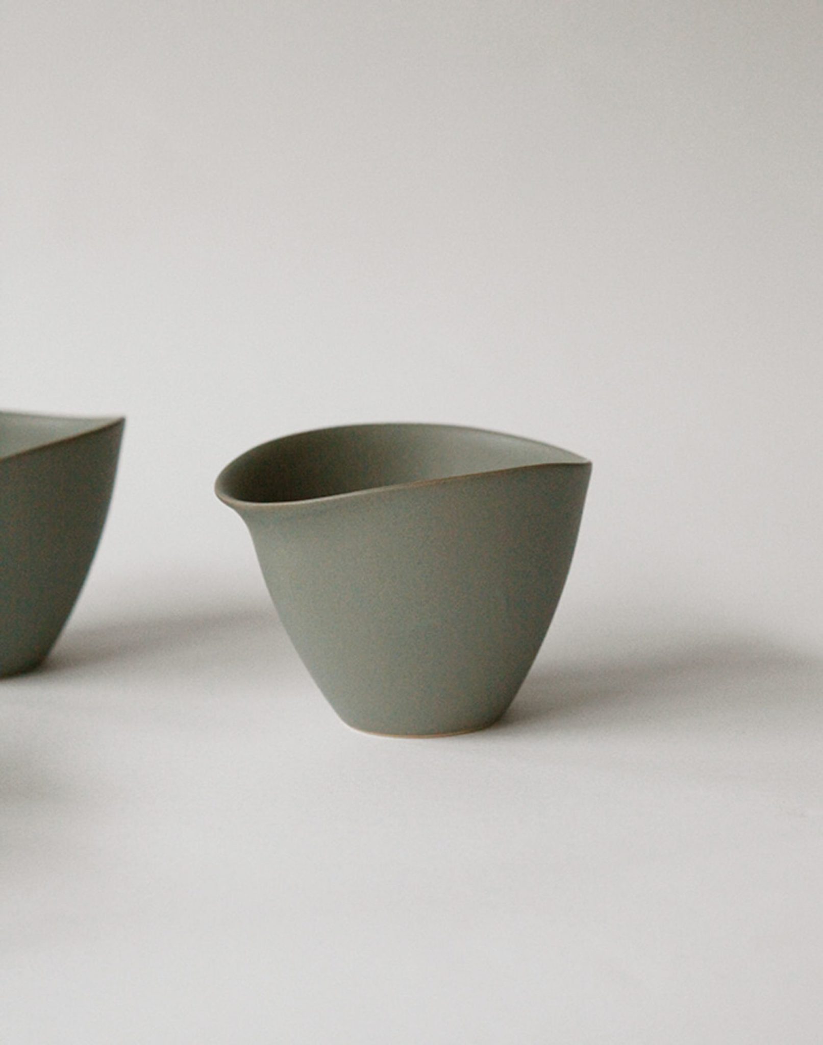 A minimalist gray NR Ceramics HIN Tea Cup with an asymmetrical wavy rim and ergonomic design rests on a white surface, while another NR Ceramics HIN cup is partially visible on the left.