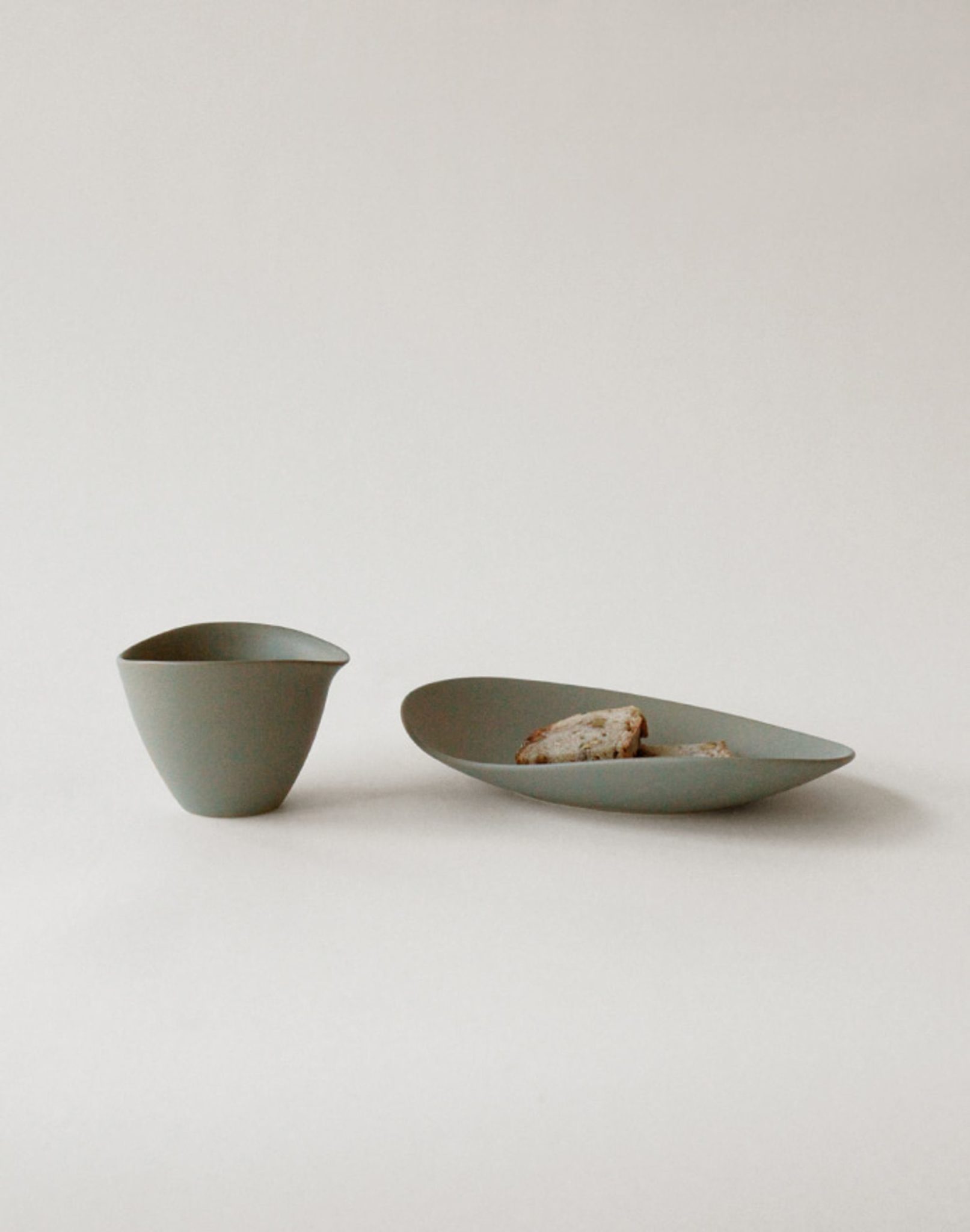 A minimalist scene with the NR Ceramics HIN Tea Cup and matching gray oval plate, both by NR Ceramics, holding two pieces of bread against a plain light background.