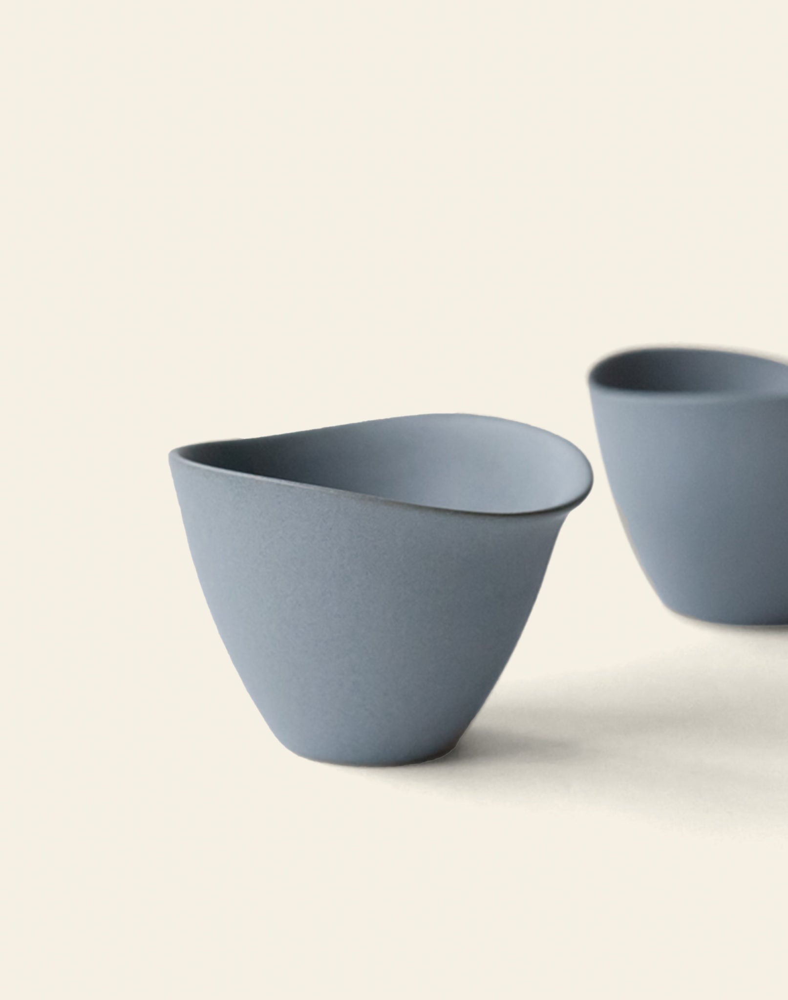 Two matte gray NR Ceramics HIN Tea Cups with smooth, curved edges display ergonomic design on a light background—one in front and one partly behind—capturing the minimalist essence of the NR Ceramics collection.
