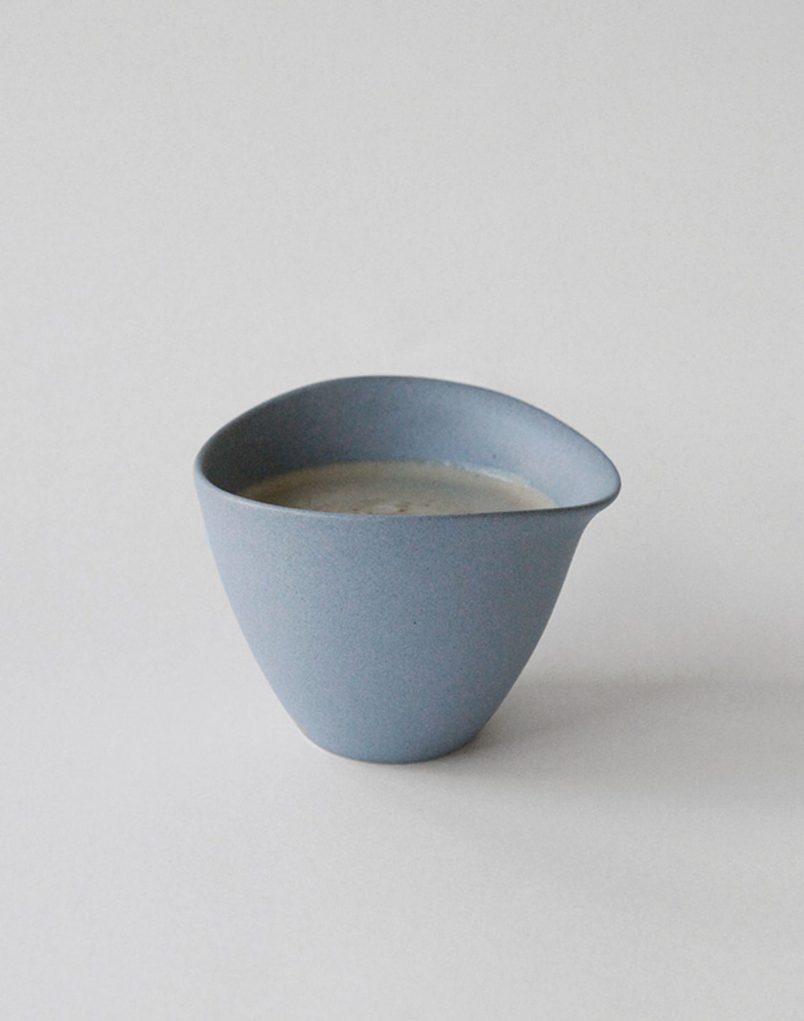 The NR Ceramics HIN Tea Cup by NR Ceramics features a minimalist, ergonomic design with an asymmetrical rim, shown filled with a creamy liquid against a plain light background.