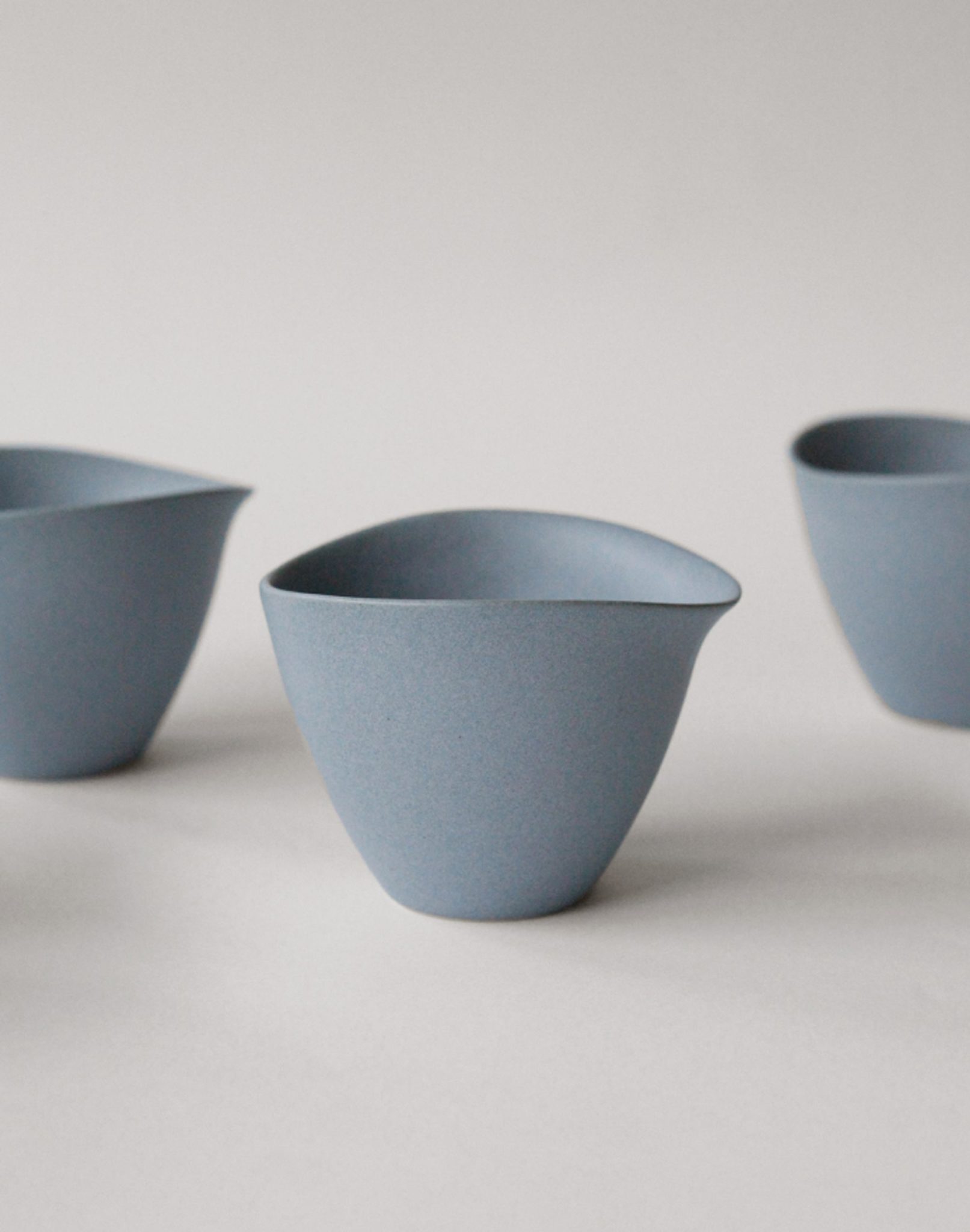 Three matte blue NR Ceramics HIN Tea Cups with wide, asymmetrical rims are arranged on a light surface against a plain white background, showcasing their minimalist modern style and ergonomic design for comfortable use.