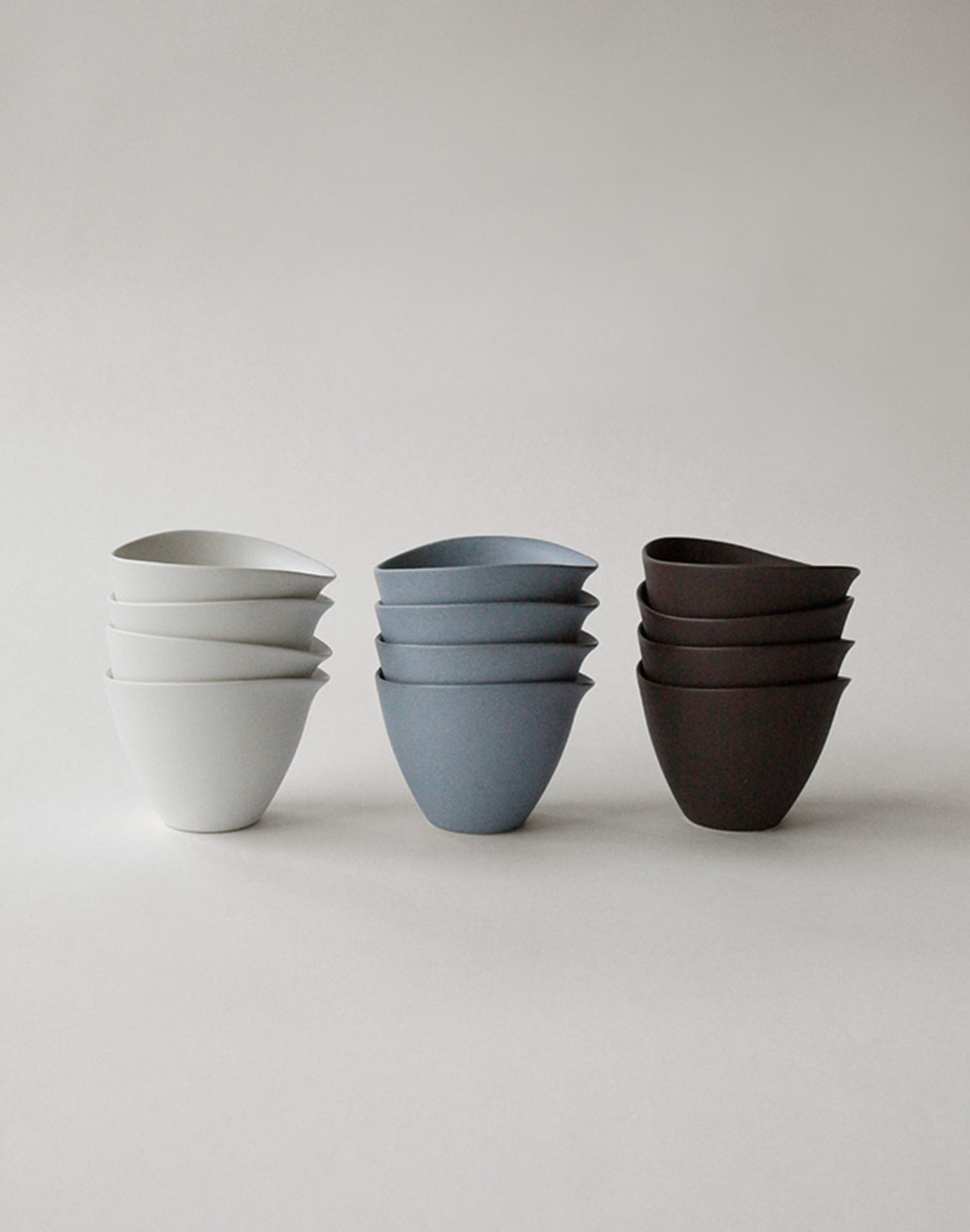 Three stacks of NR Ceramics HIN Tea Cups by NR Ceramics on a neutral background: four white on the left, four blue in the center, and four dark brown on the right—all with ergonomic designs for comfort.