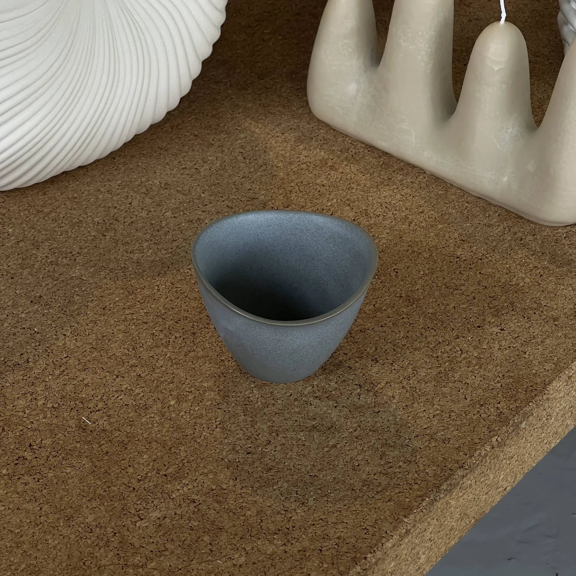A compact, matte gray NR Ceramics HIN Tea Cup with an ergonomic design rests on a cork surface, accompanied by abstract white ceramic decor in the background.