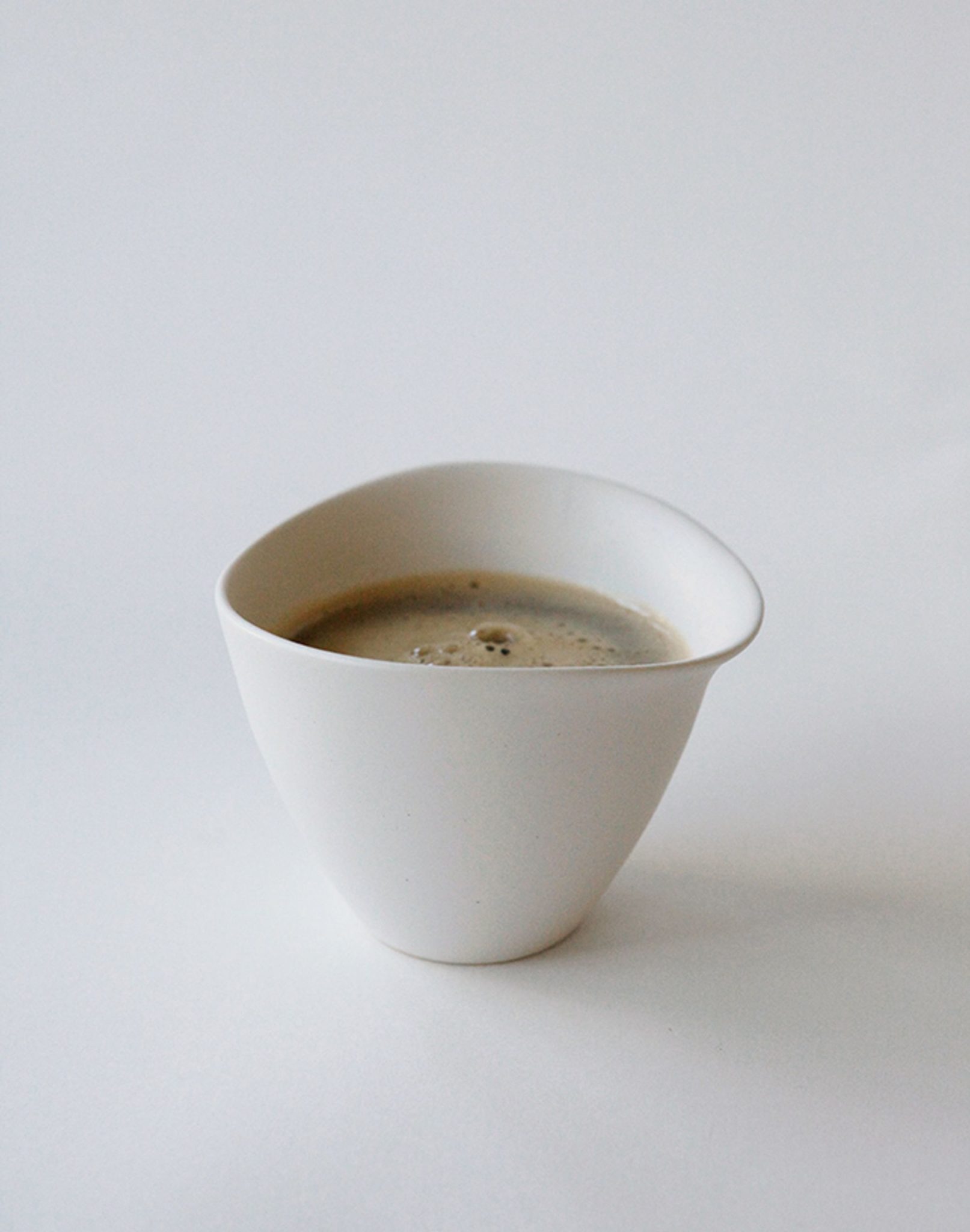 The NR Ceramics HIN Tea Cup by NR Ceramics is a white ceramic cup with an ergonomic design, shown filled with coffee on a plain white background.