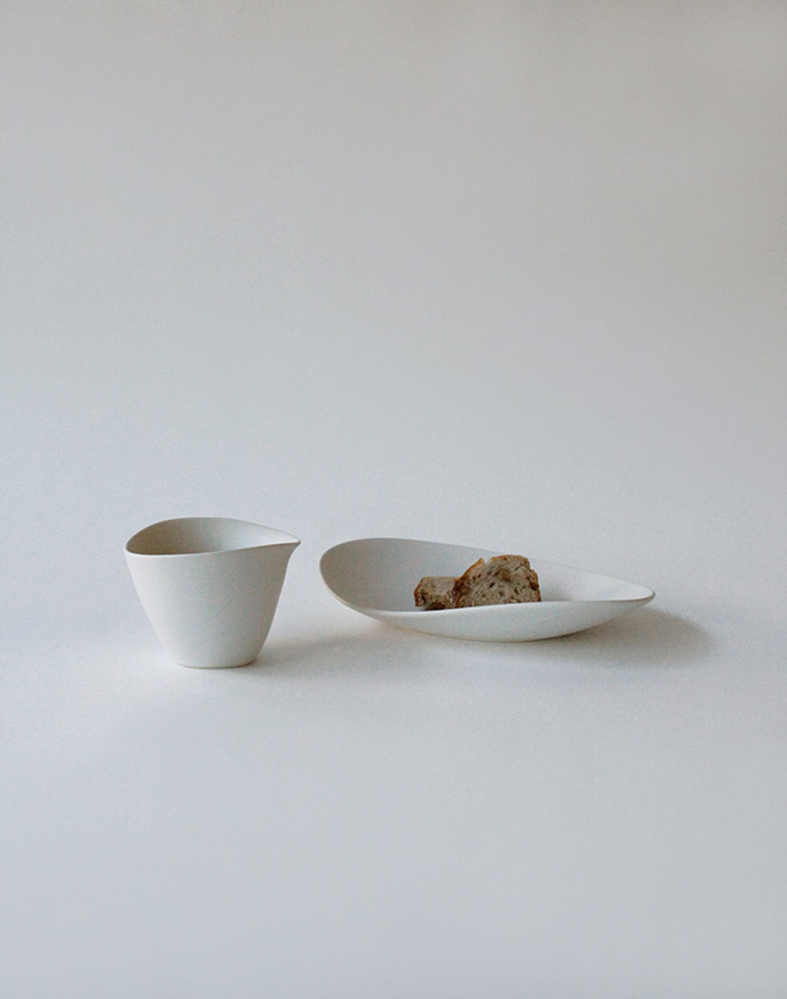 A minimalist scene featuring the NR Ceramics HIN Tea Cup and a white shallow bowl with a small piece of brown bread, all set against a plain light background.