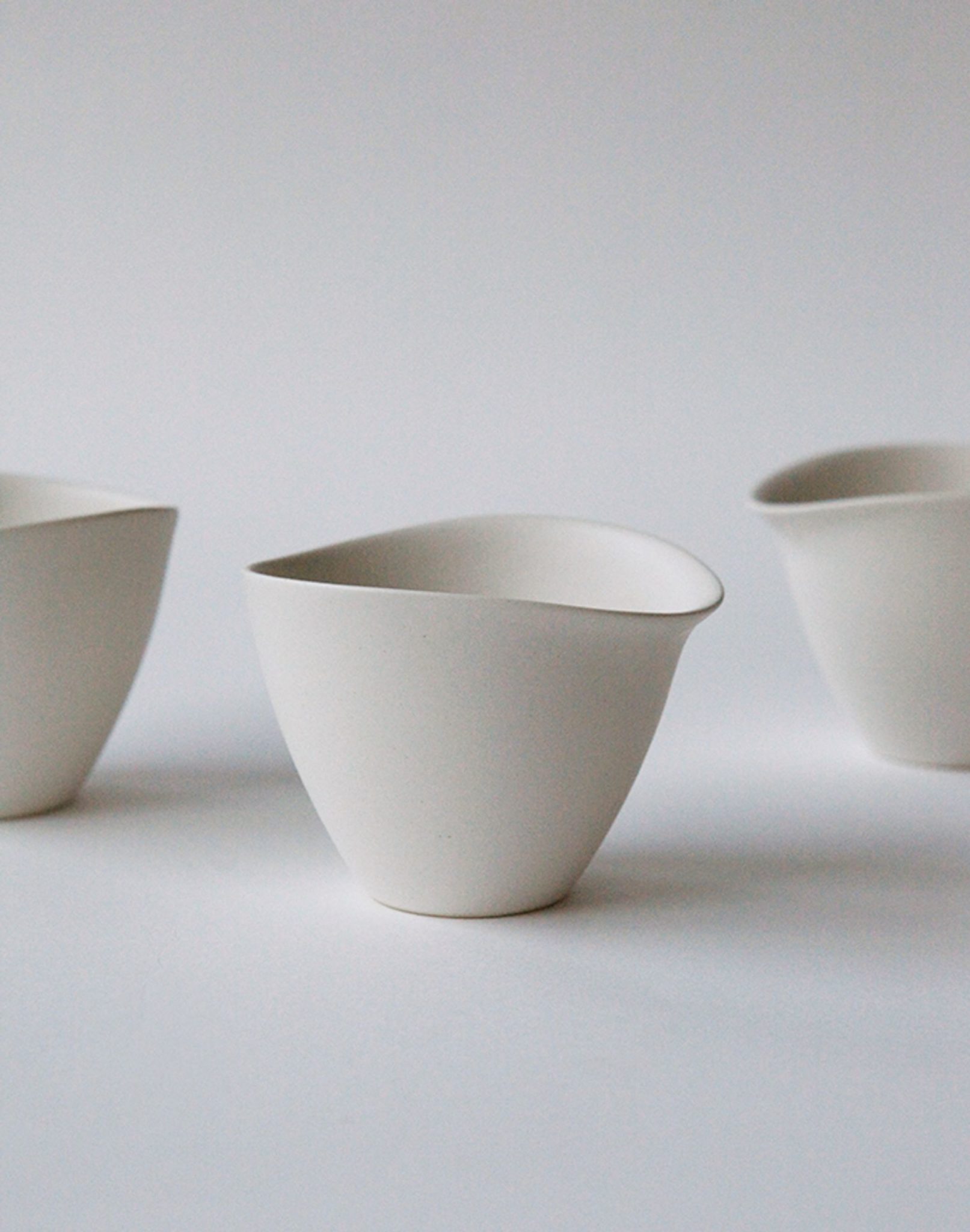 Three minimalist, white NR Ceramics HIN Tea Cups with irregular, organic shapes are arranged on a plain, light background. Their ergonomic design and thoughtful spacing create a clean, modern composition.