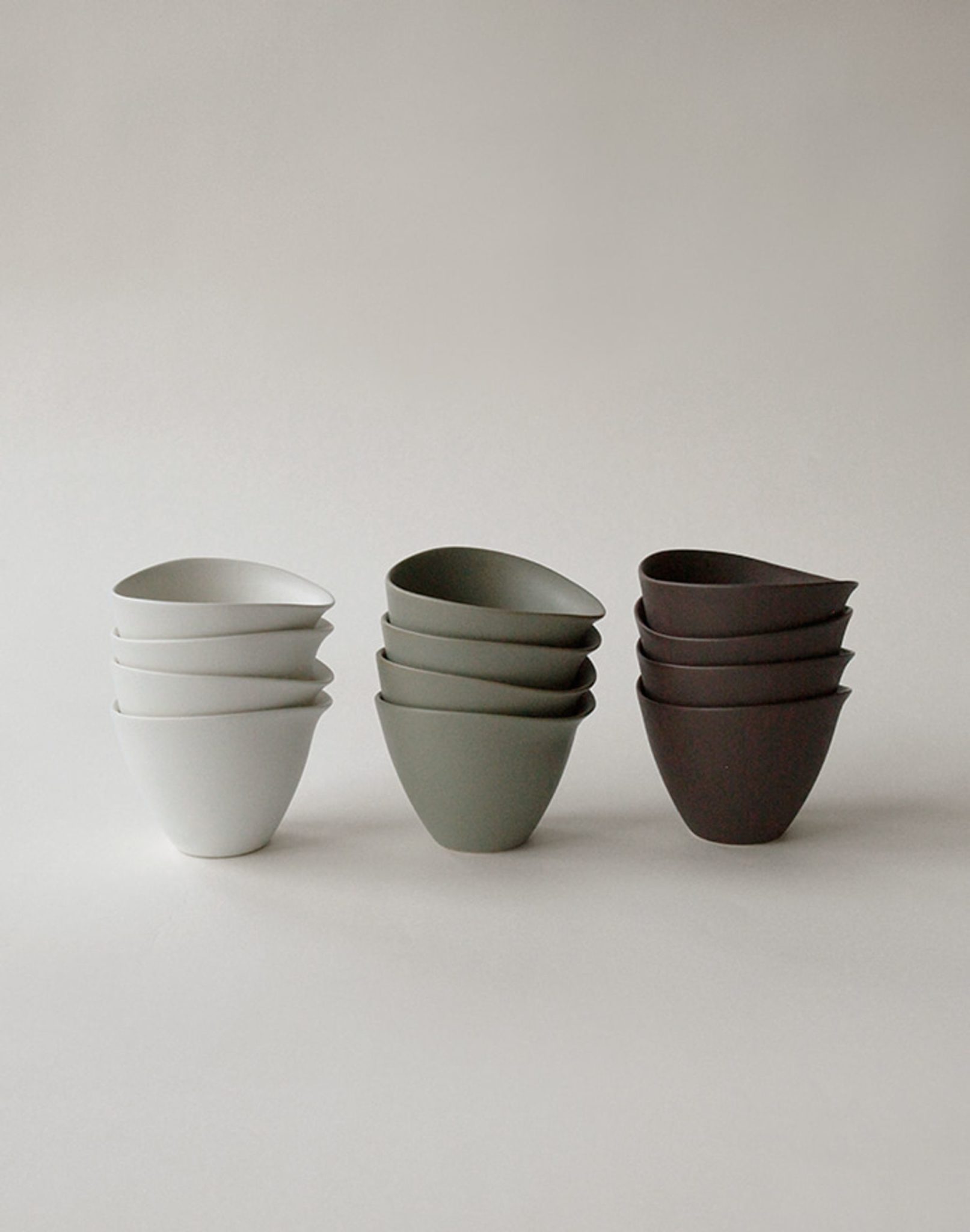 Three stacks of NR Ceramics HIN Tea Cups by NR Ceramics in white, sage green, and dark brown are arranged side by side against a plain background. Each stack has three minimalist, handleless cups with an ergonomic design nested together.