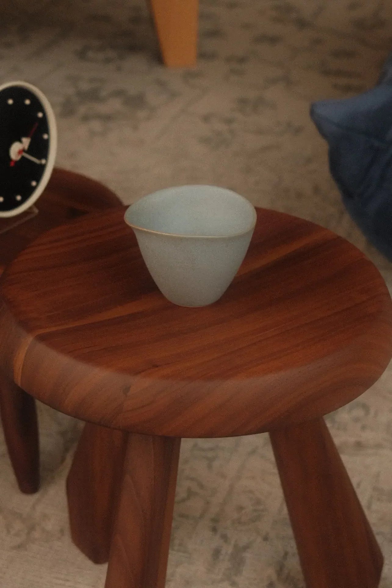 A light blue NR Ceramics HIN Tea Cup with an ergonomic design rests on a smooth round wooden stool; a clock and cushioned surface appear in the background.