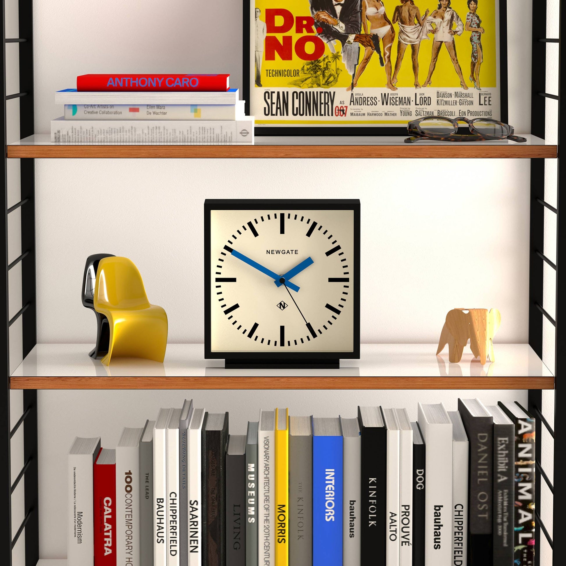 A bookshelf styled with books, decor, the Newgate Amp Mantel Clock by Newgate, a yellow abstract sculpture, a wooden elephant, and a Dr. No movie poster on the top shelf brings modern flair to your space.