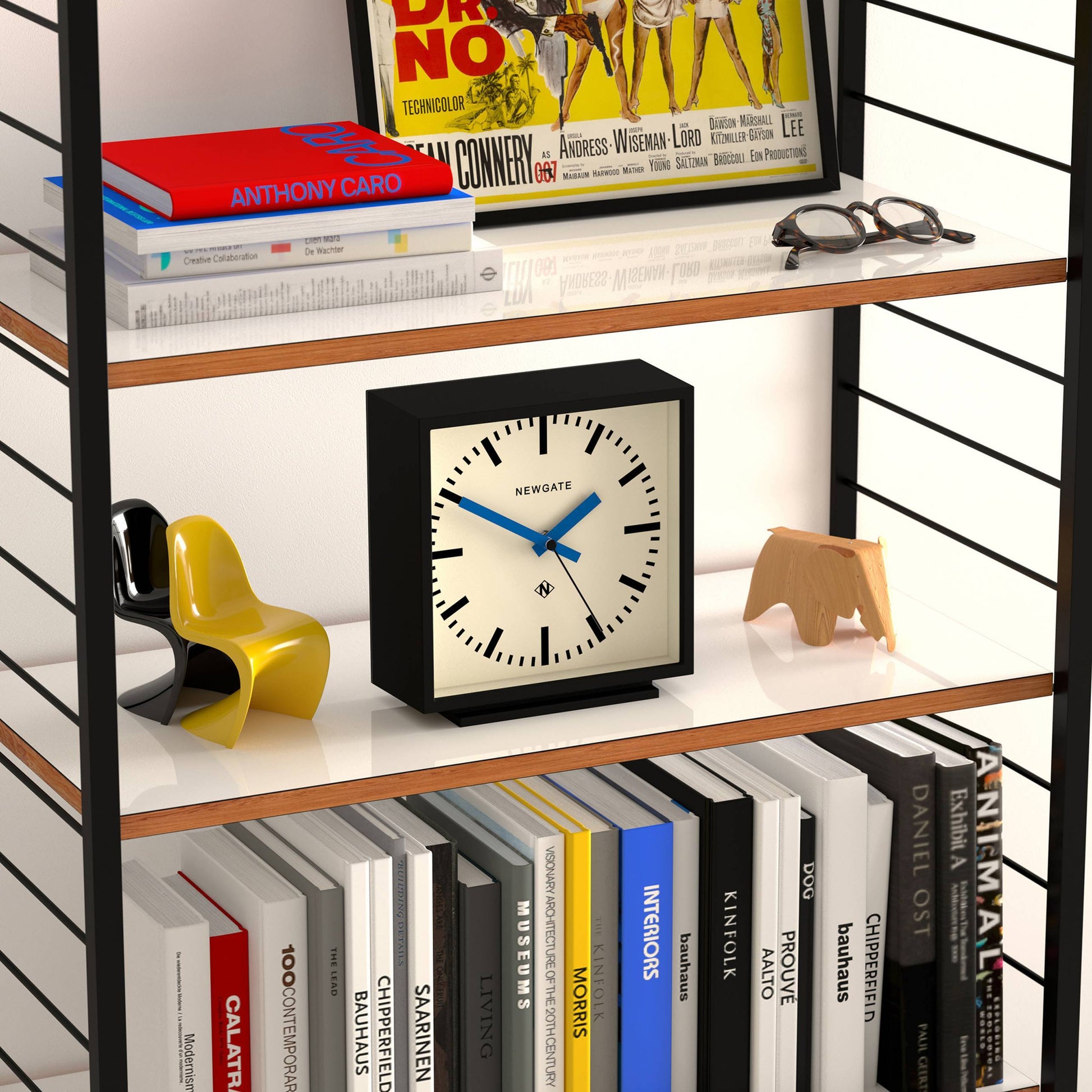 The Newgate Amp Mantel Clock by Newgate, featuring a cream face and black frame, sits on a shelf between a yellow chair, wooden animal figurine, and books, combining classic station style with modern flair among records and magazines above.