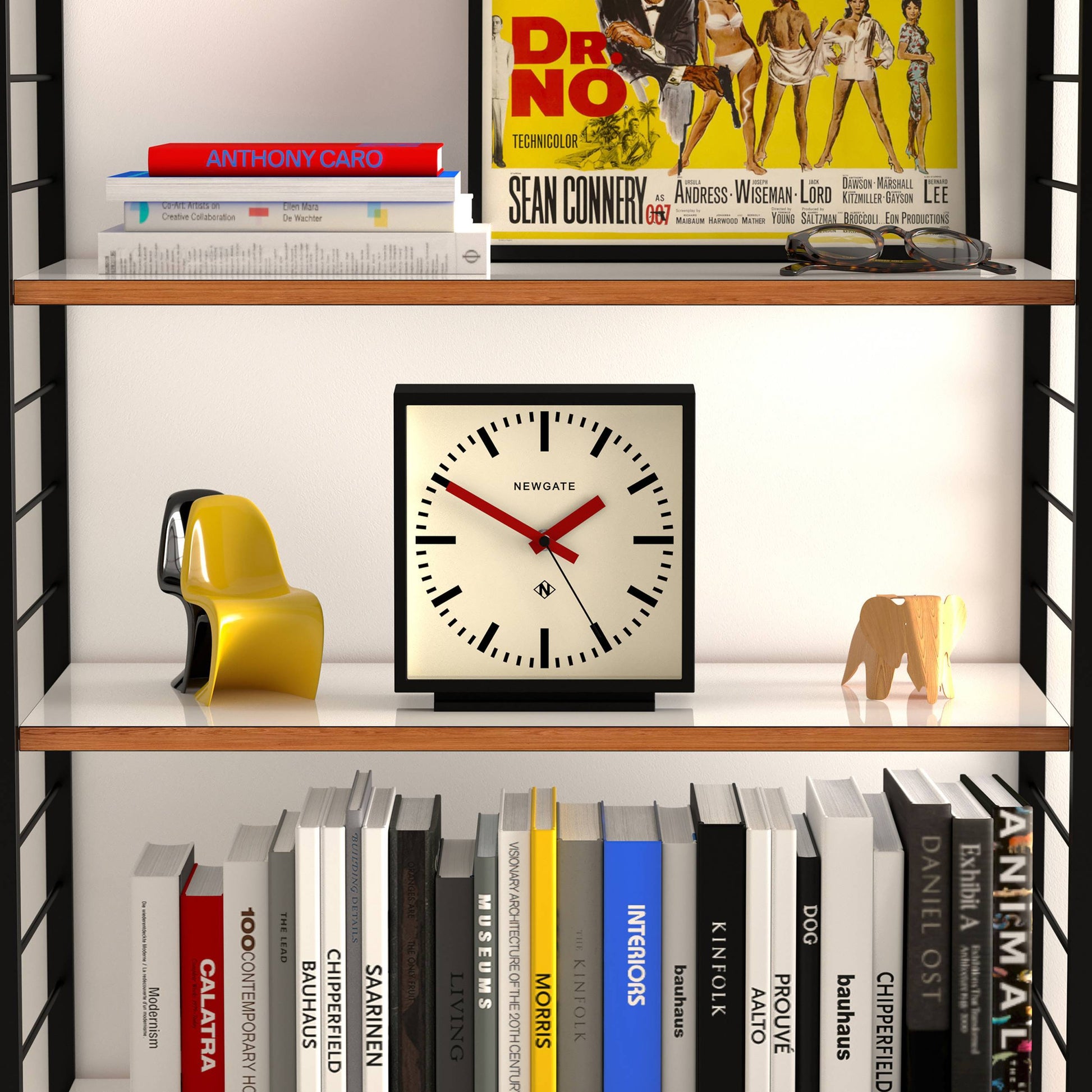 A bookshelf displays a Newgate Amp Mantel Clock, books, a yellow abstract sculpture, and an elephant figurine. The top shelf features a vintage Dr. No movie poster and an Anthony Caro book for modern style.