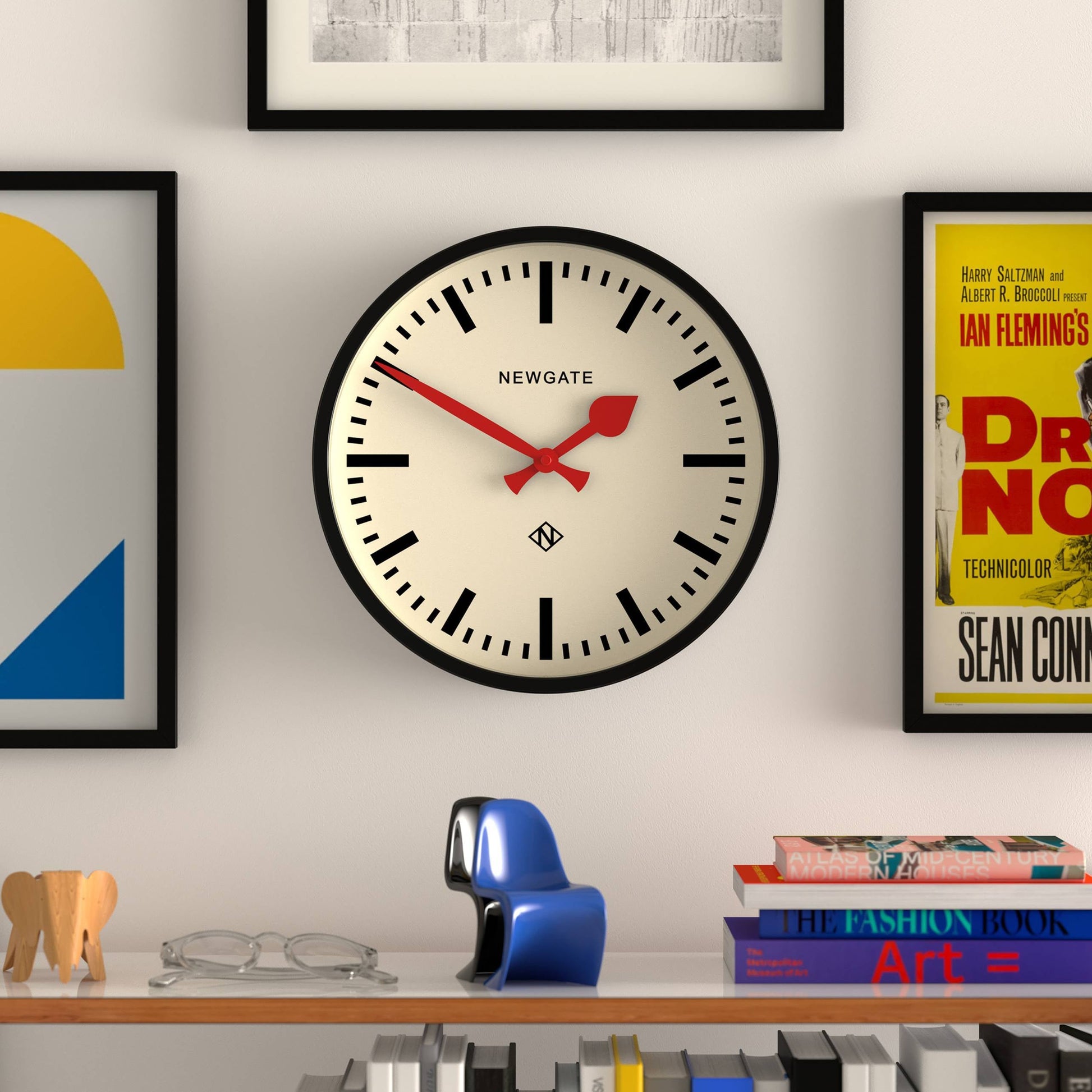 A Newgate Luggage Wall Clock by Newgate, with black hour marks and red hands, is displayed on a white wall among framed art. A shelf beneath holds books, a blue figurine, and wooden animal decorations.