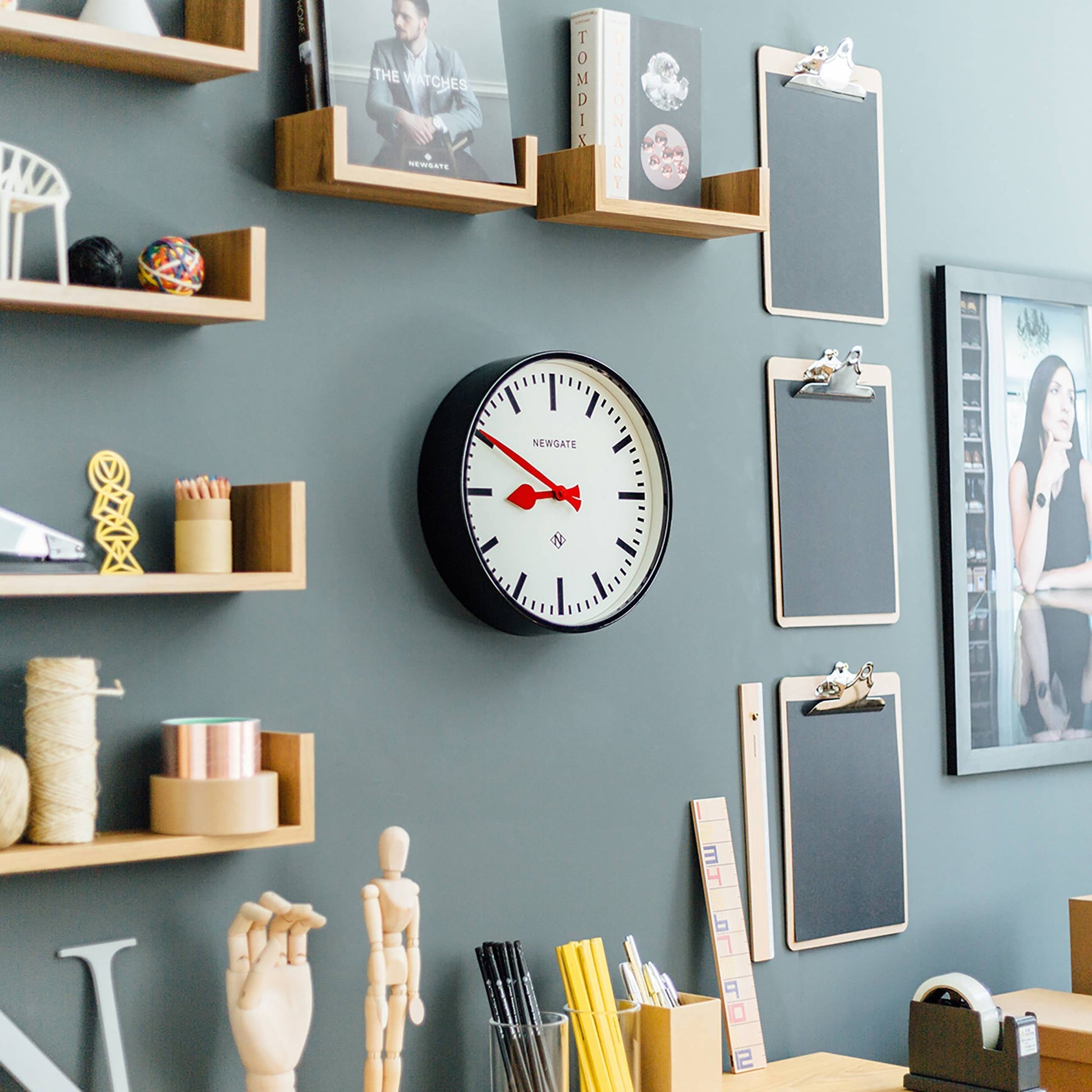 A modern office wall displays a Newgate Luggage Wall Clock by Newgate for retro charm, floating wooden shelves with books and décor, three blank clipboards, and various stationery items arranged on the desk below.