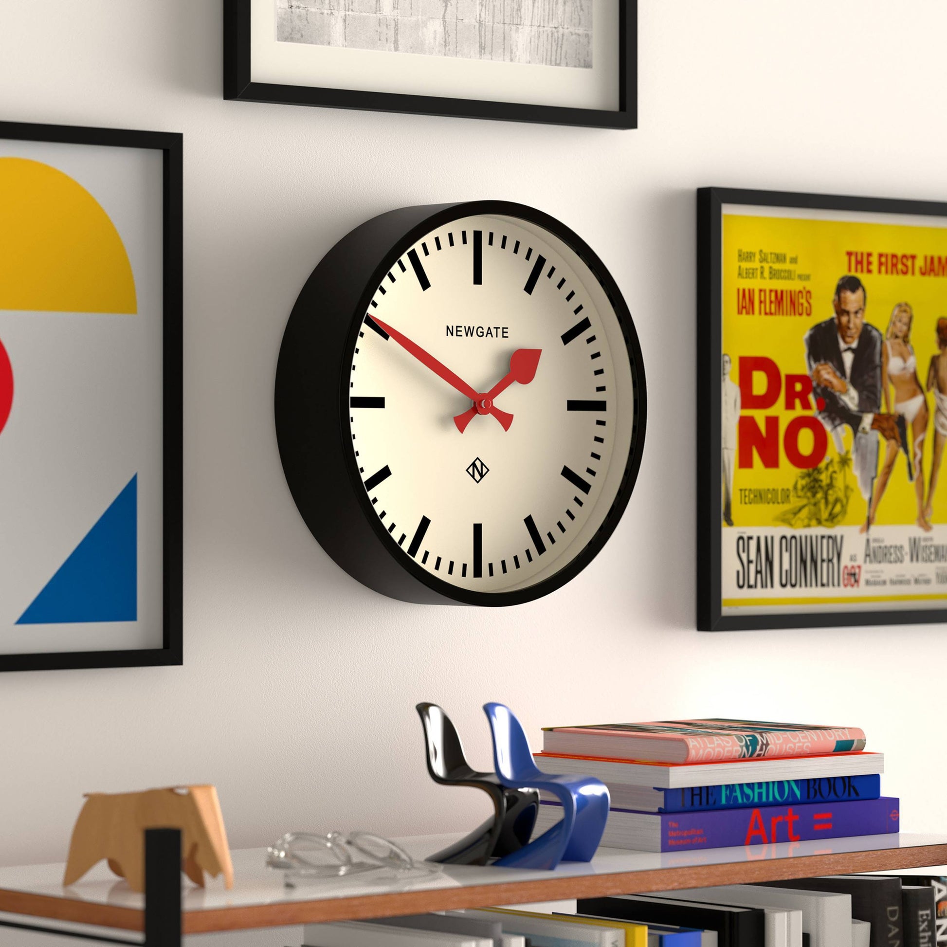 A Newgate Luggage Wall Clock by Newgate with a black frame and red hands hangs on a white wall, surrounded by colorful art prints and posters, above a shelf holding books and decor.