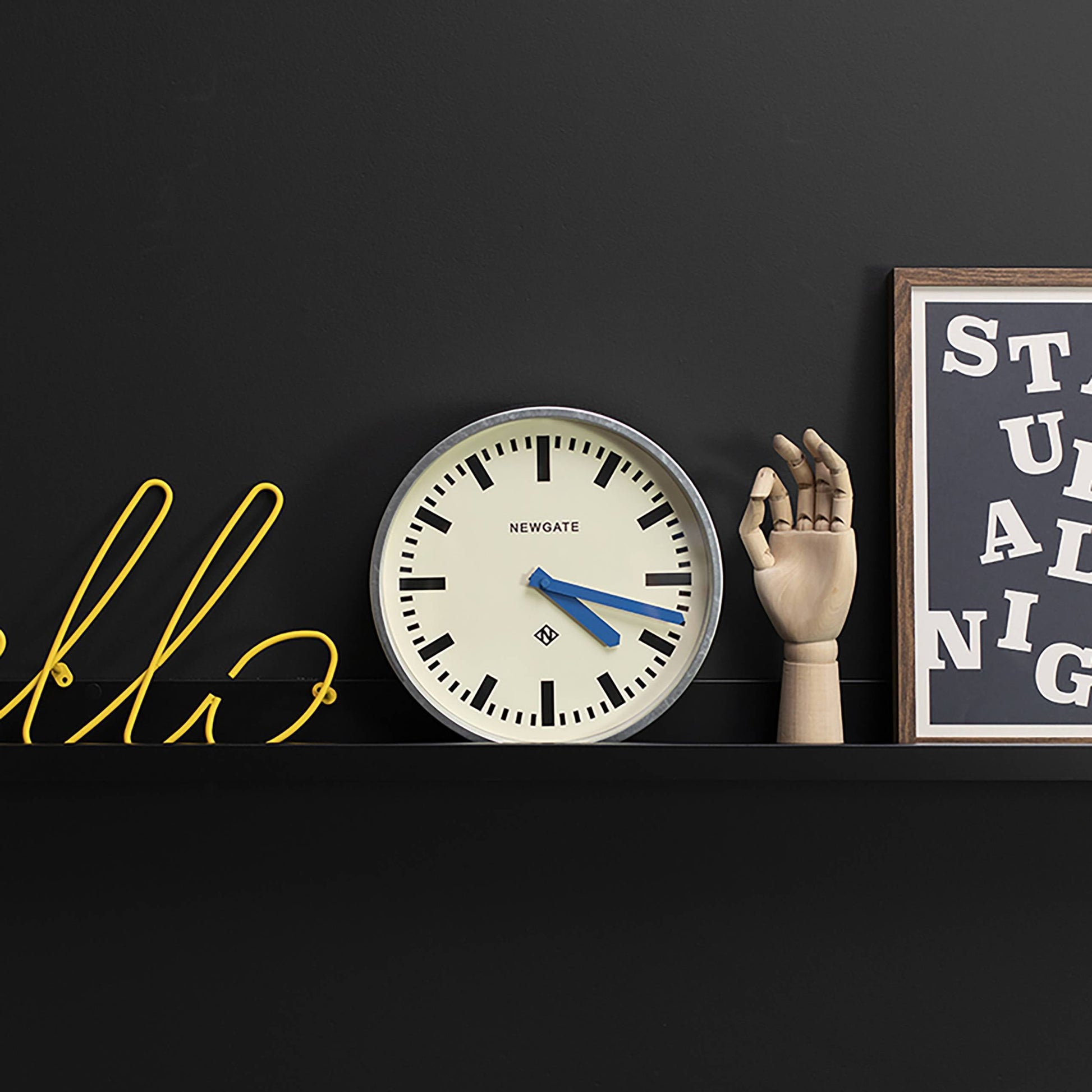 A minimalist black wall shelf displays a yellow wire word in cursive, a Newgate Luggage Wall Clock Galvanised by Newgate with blue hands, an upright wooden mannequin hand, and framed abstract black-and-white typographic art.