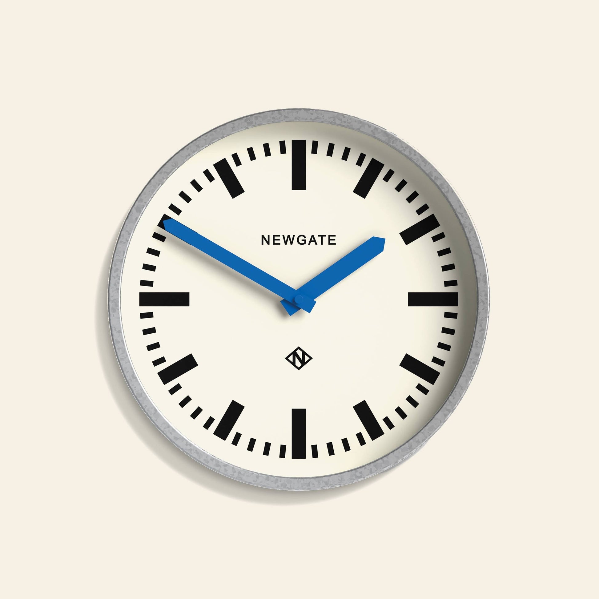 The Newgate Luggage Wall Clock Galvanised features a round white face, black markings, and bold blue hands set to 10:10. Reminiscent of mid-century railway clocks, “Newgate” appears above the center.