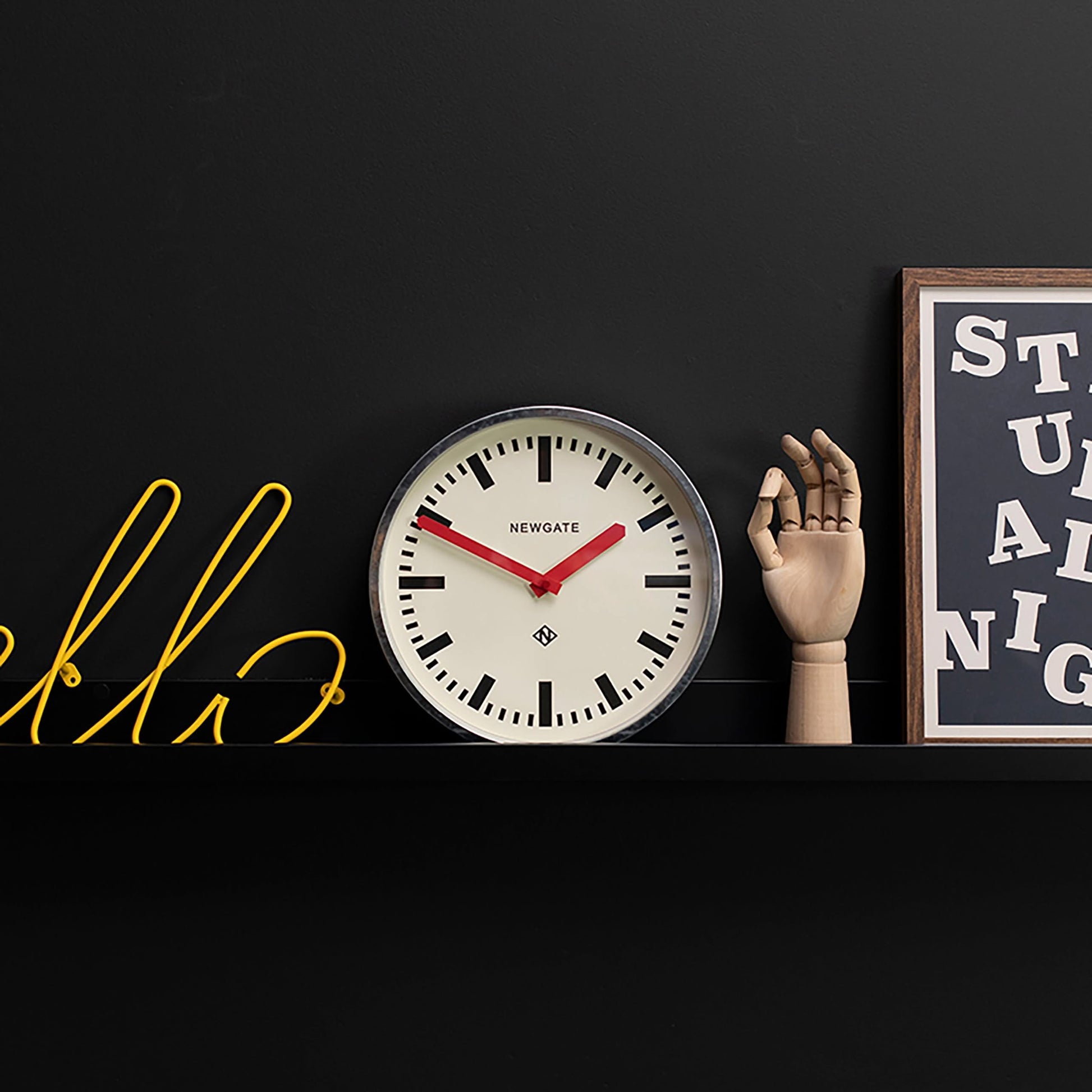 The Newgate Luggage Wall Clock Galvanised, featuring red hands and an industrial look inspired by mid-century railway clocks, rests on a black shelf beside a yellow neon hello sign, a wooden mannequin hand, and a framed white-on-black print.