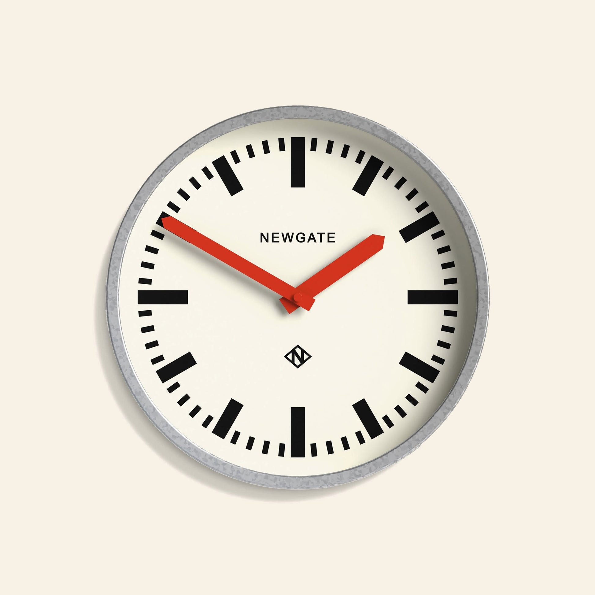 The Newgate Luggage Wall Clock Galvanised features a round, industrial design with a white face, bold black numerals, red hands, and NEWGATE branding at the top. Inspired by mid-century railway clocks, it displays the time 10:10.