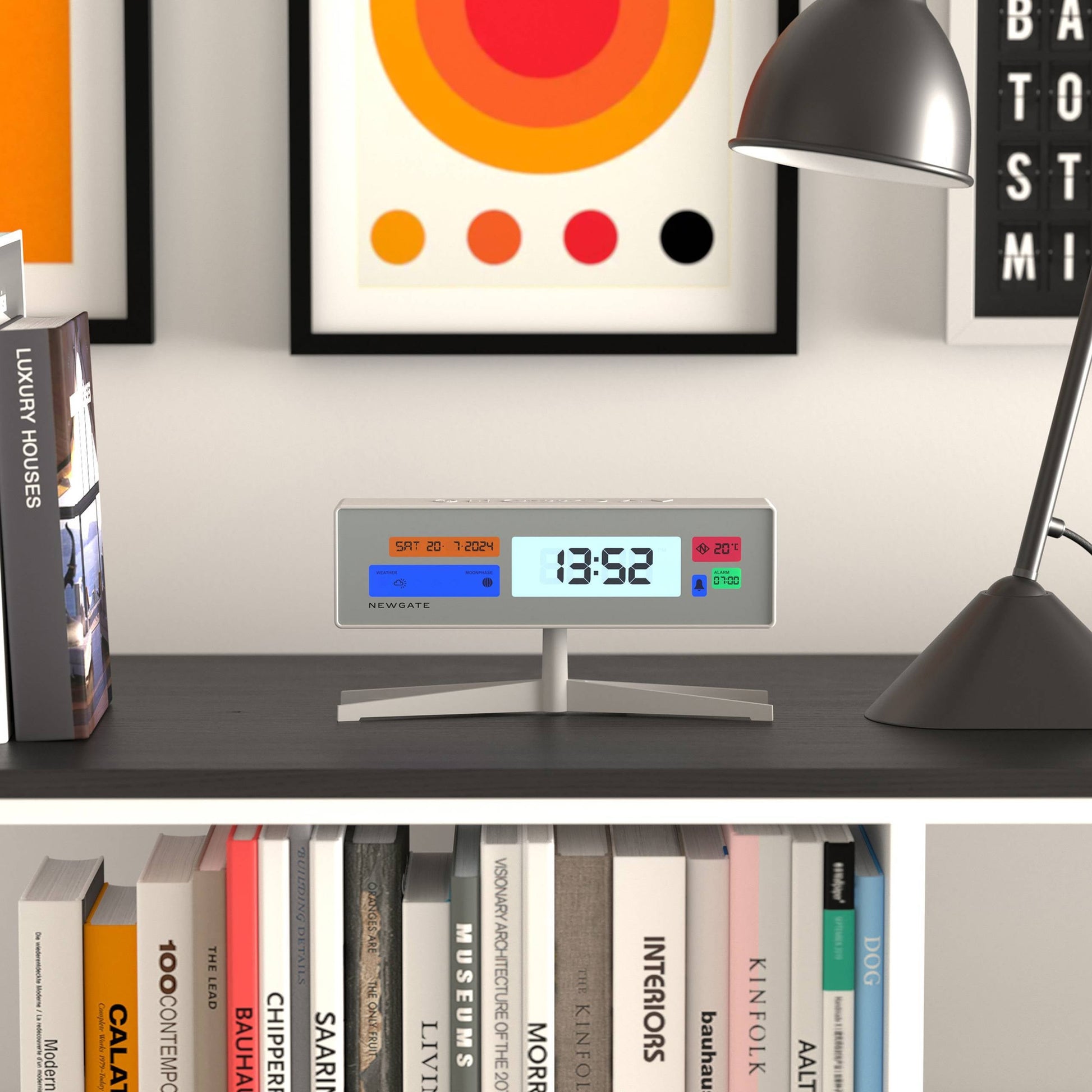 The Newgate Supergenius LCD Alarm Clock by Newgate displays 13:52 and temperature as it rests on a dark shelf beside a black desk lamp, books, and vibrant artwork; its USB cable keeps it connected for convenience.