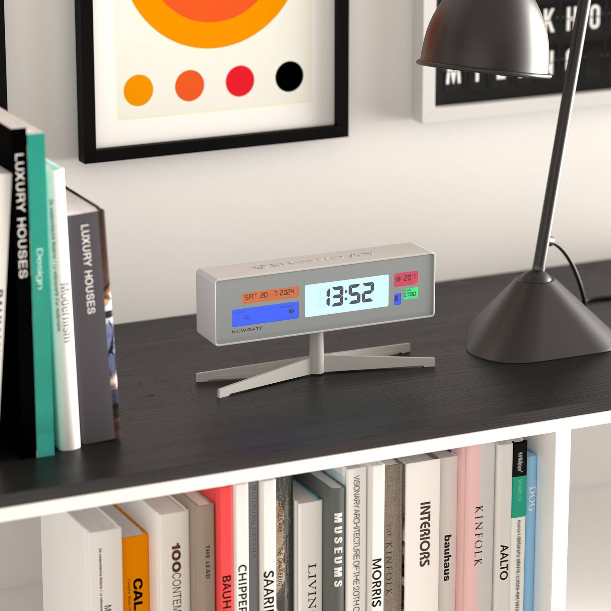 The Newgate Supergenius LCD Alarm Clock by Newgate displays 13:52 as it sits on a black desk near a black lamp, surrounded by books and colorful wall art, powered conveniently by a USB cable.