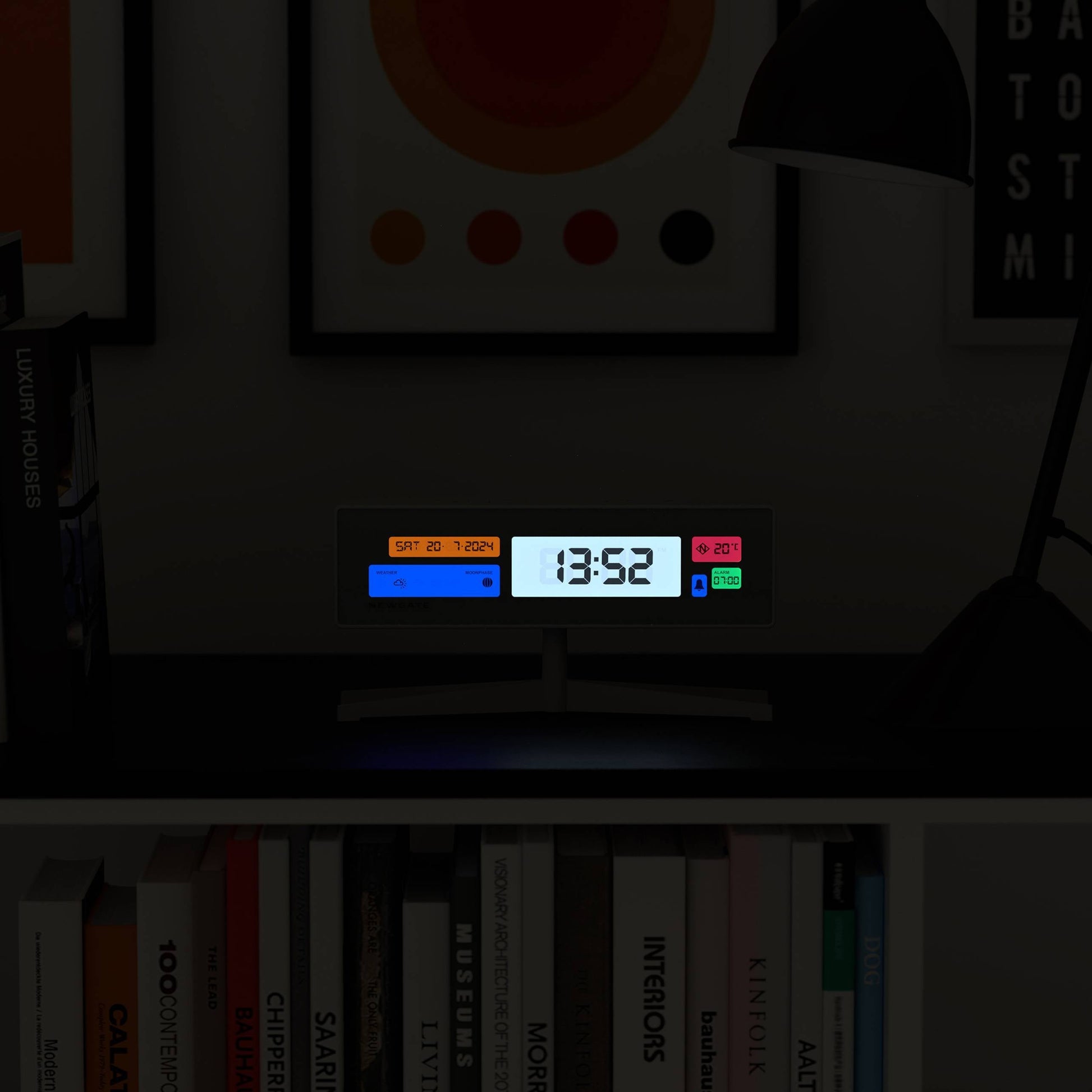 A Newgate Supergenius LCD Alarm Clock by Newgate, showing 13:52, rests on a dark shelf among books and glows under a desk lamp. Framed artwork hangs behind it, while a USB cable powers the clock.