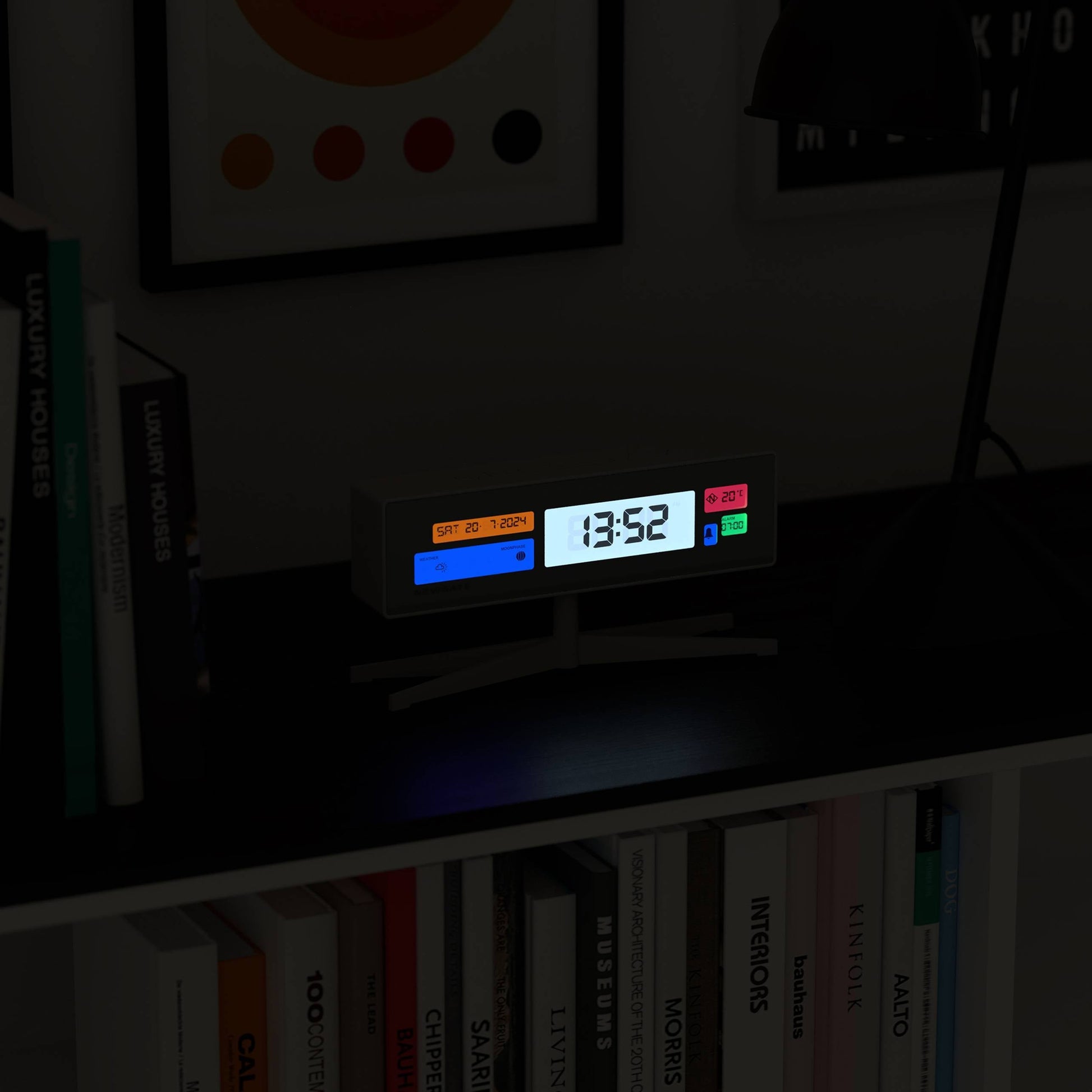 The Newgate Supergenius LCD Alarm Clock sits on a black shelf beside books and a lamp, its colorful buttons and glowing temperature display visible. A USB cable connects the clock, while framed artwork hangs above.