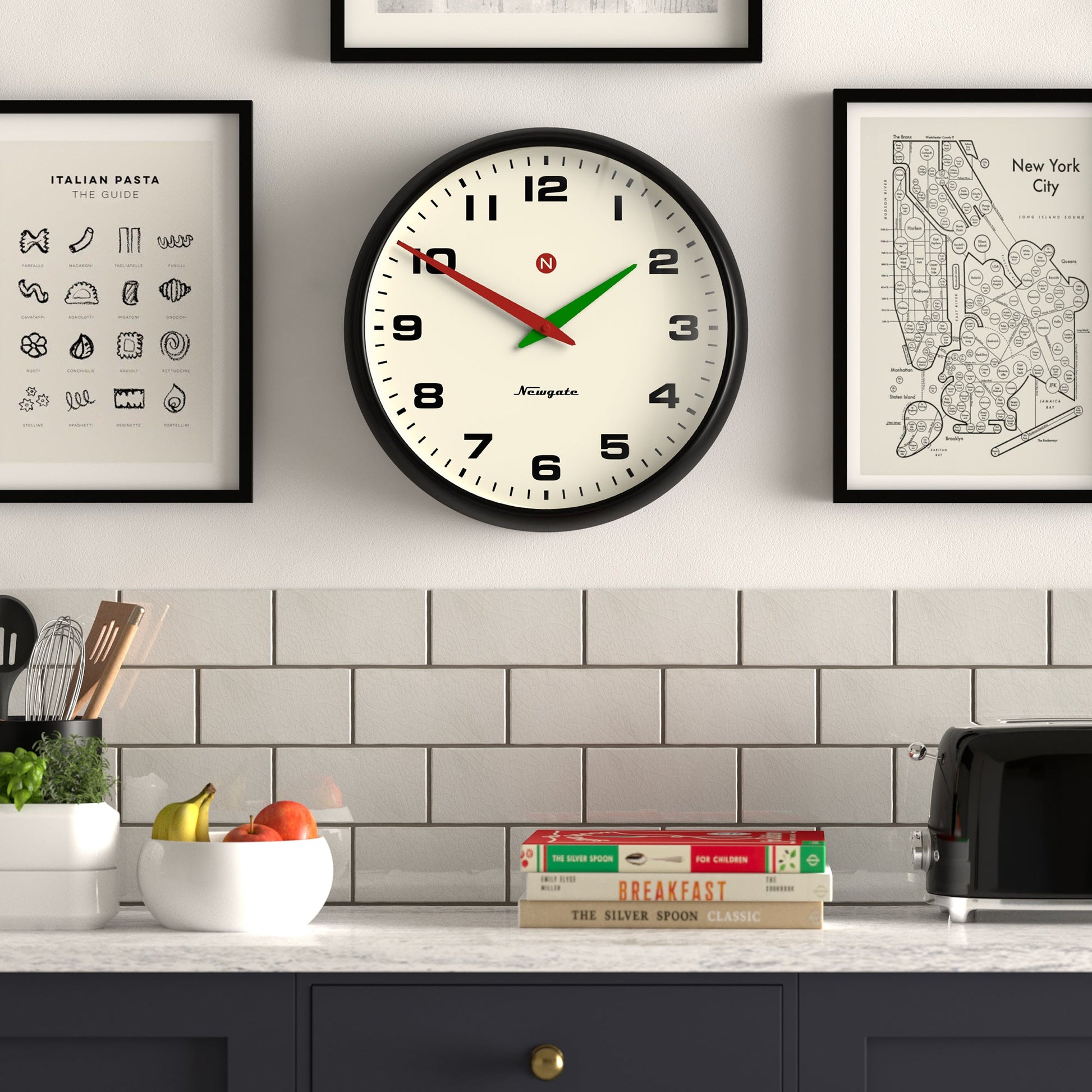 A Newgate Superstore Wall Clock with a black frame hangs above the kitchen counter, flanked by two framed prints. On the counter are a fruit bowl, two cookbooks, a toaster, and small plants in front of white subway tile.