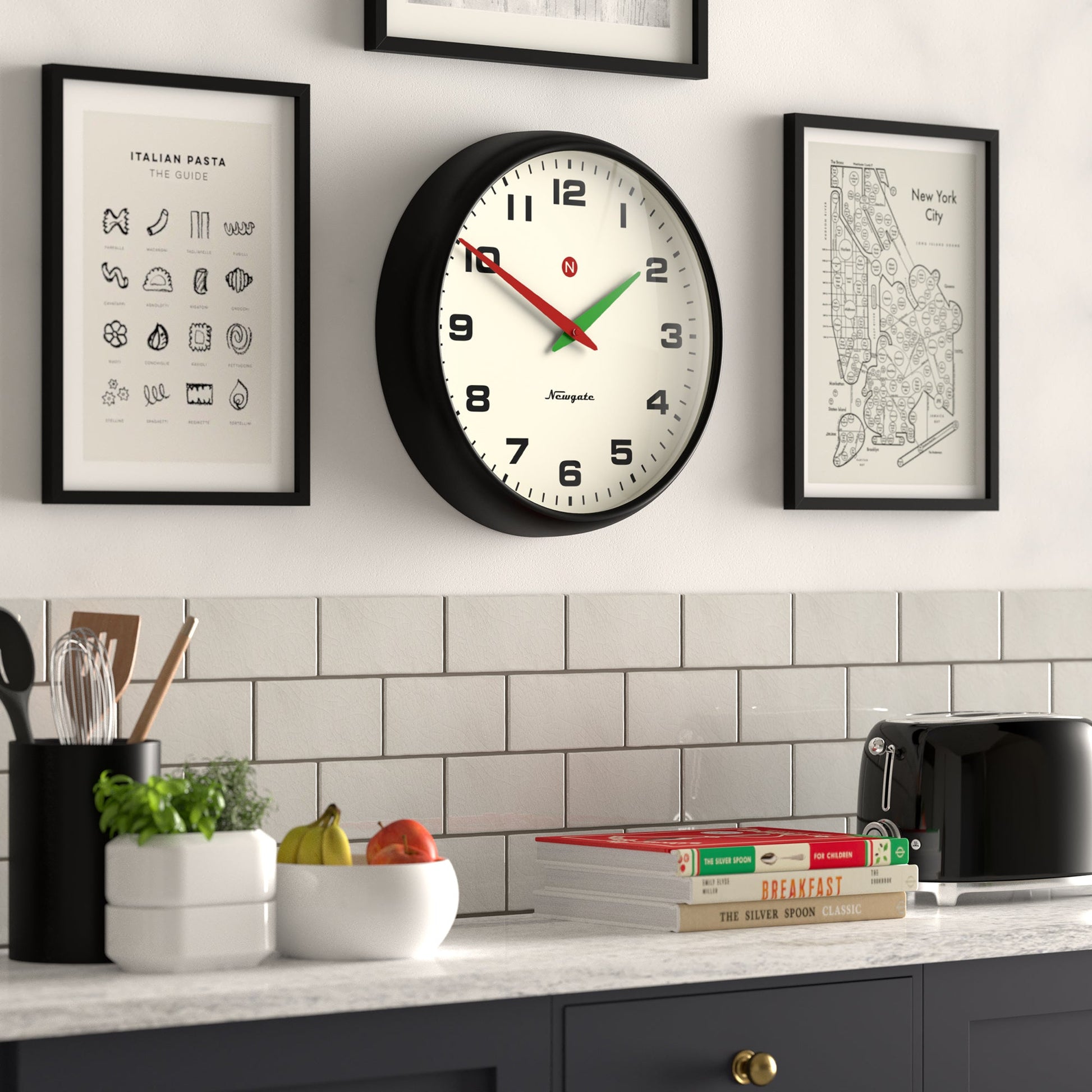 A kitchen wall is adorned with a Newgate Superstore Wall Clock by Newgate, framed art, and a white subway tile backsplash. On the counter are fruit, utensils, a toaster, and stacked cookbooks. The clock shows 2:15.