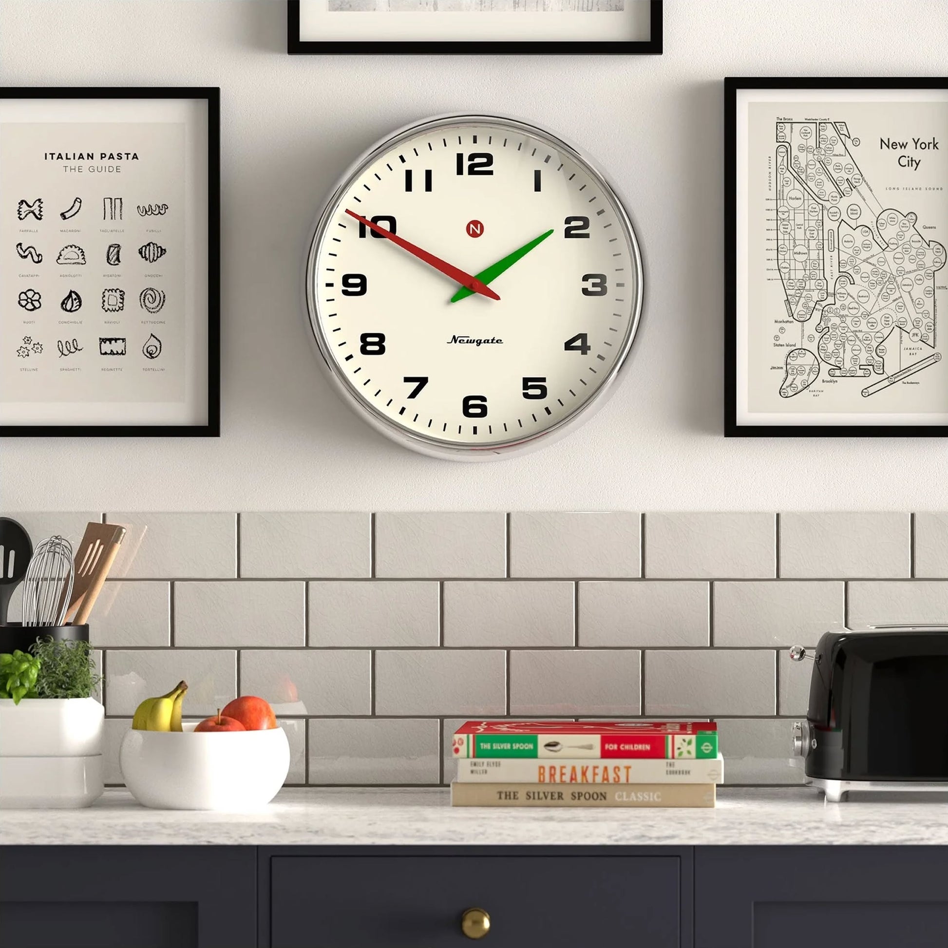 A Newgate Superstore Wall Clock by Newgate shows 10:10 above a countertop with a bowl of fruit, kitchen utensils, stacked recipe books, and a black toaster, set against framed art and a white-tiled backsplash.