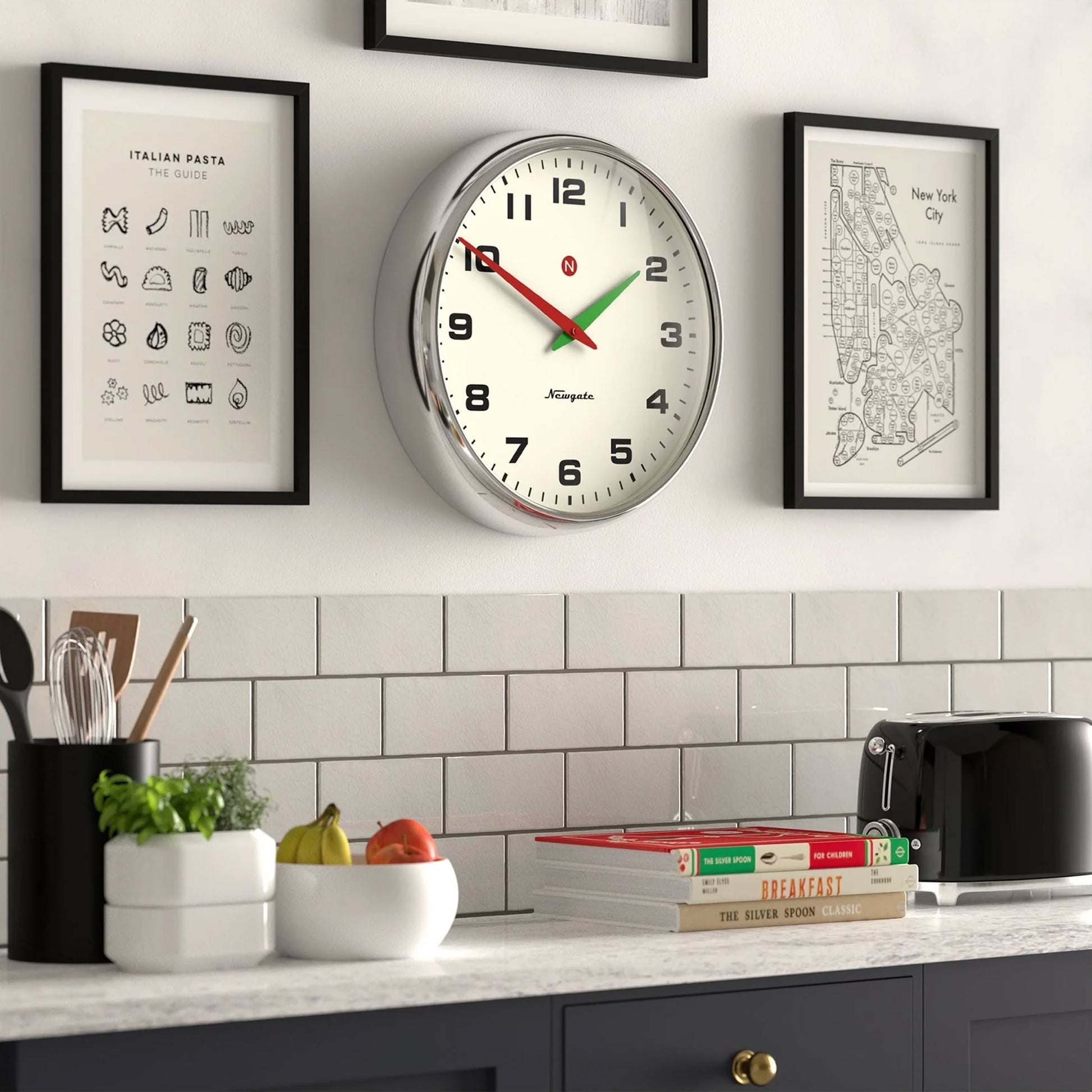 A kitchen wall features a Newgate Superstore Wall Clock showing 12:03, framed art prints, a countertop with fruit, utensils, a toaster, jars, stacked cookbooks, and a white subway tile backsplash.