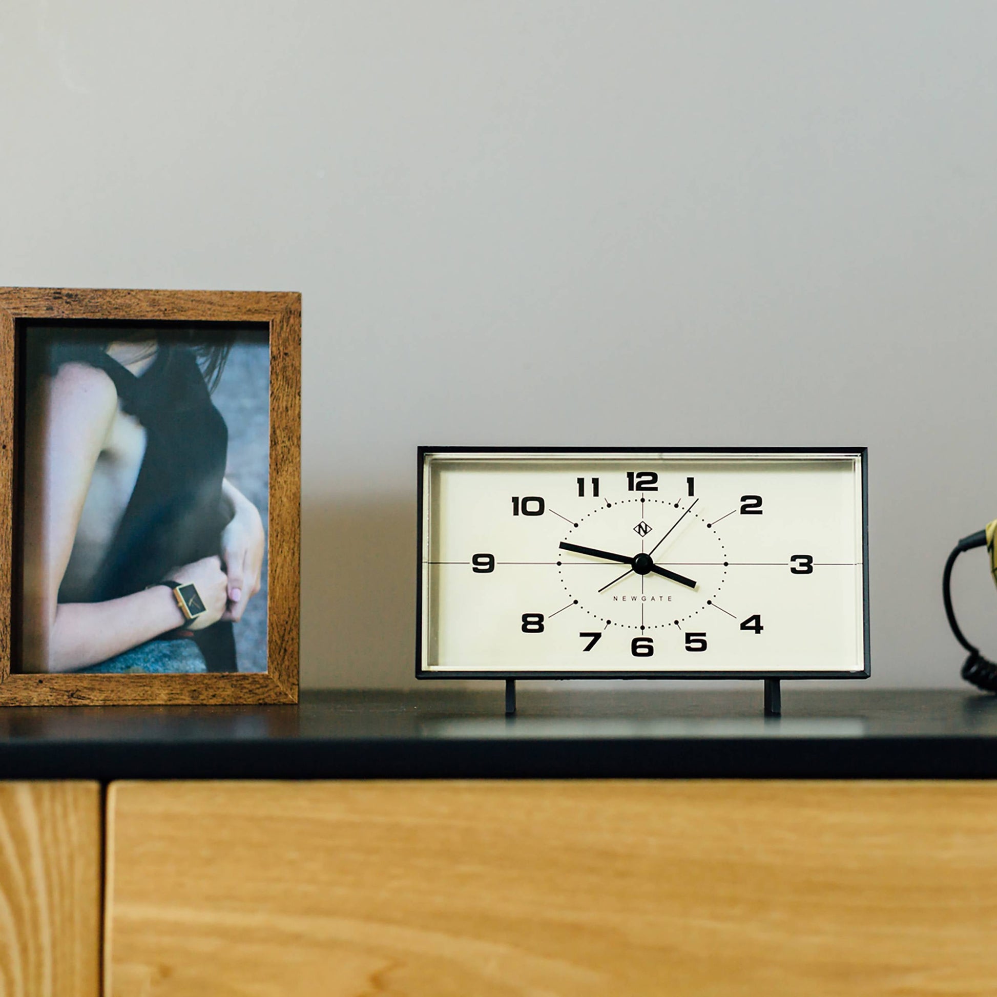 A framed photo of a woman, the Newgate Wideboy Alarm Clock by Newgate showing 10:11 with a mid-century atomic dial, and a coiled cord sit on a wooden surface against a light gray wall.