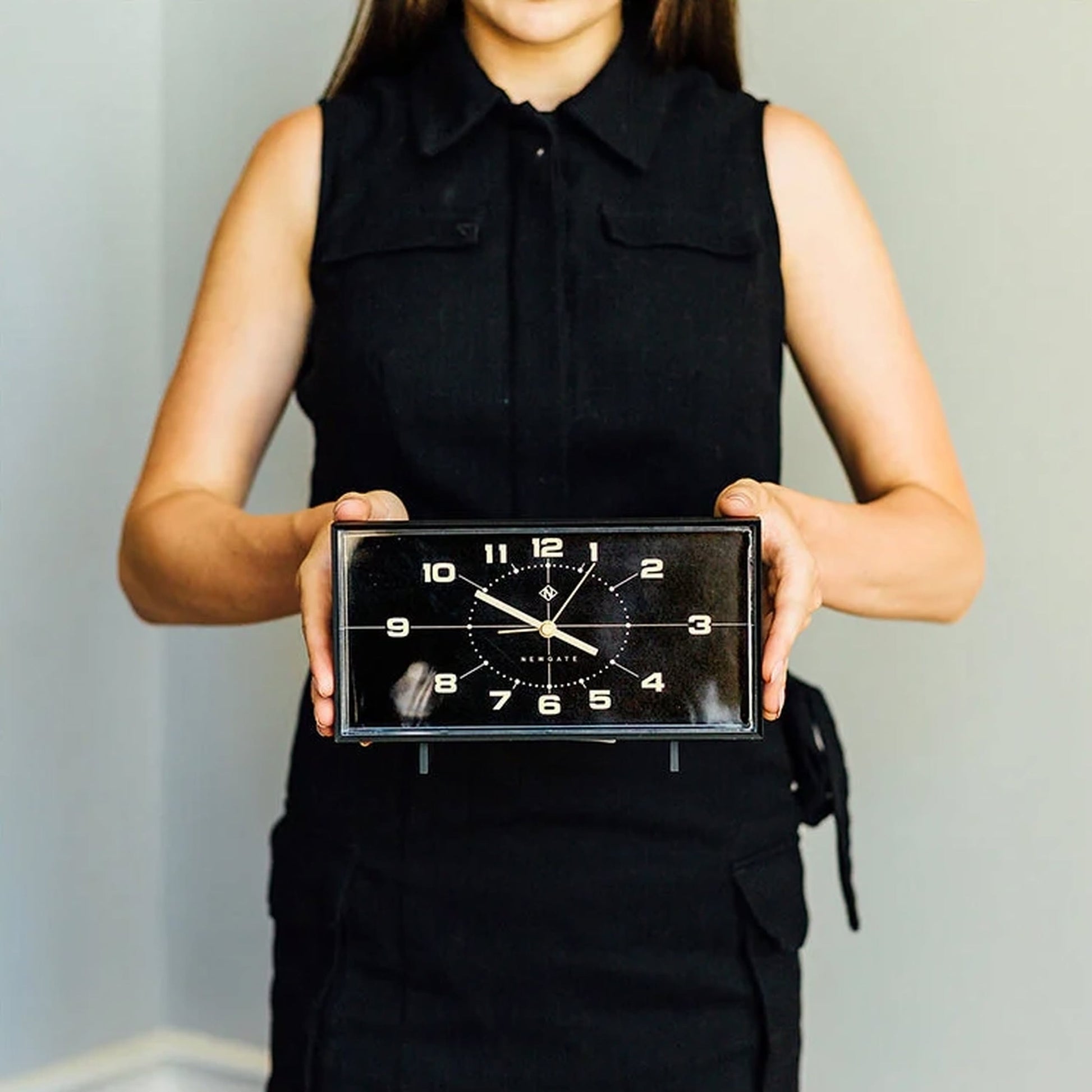 A woman in a sleeveless black dress holds the Newgate Wideboy Alarm Clock by Newgate, featuring a rectangular retro design with white numbers and hands, standing against a plain light background.