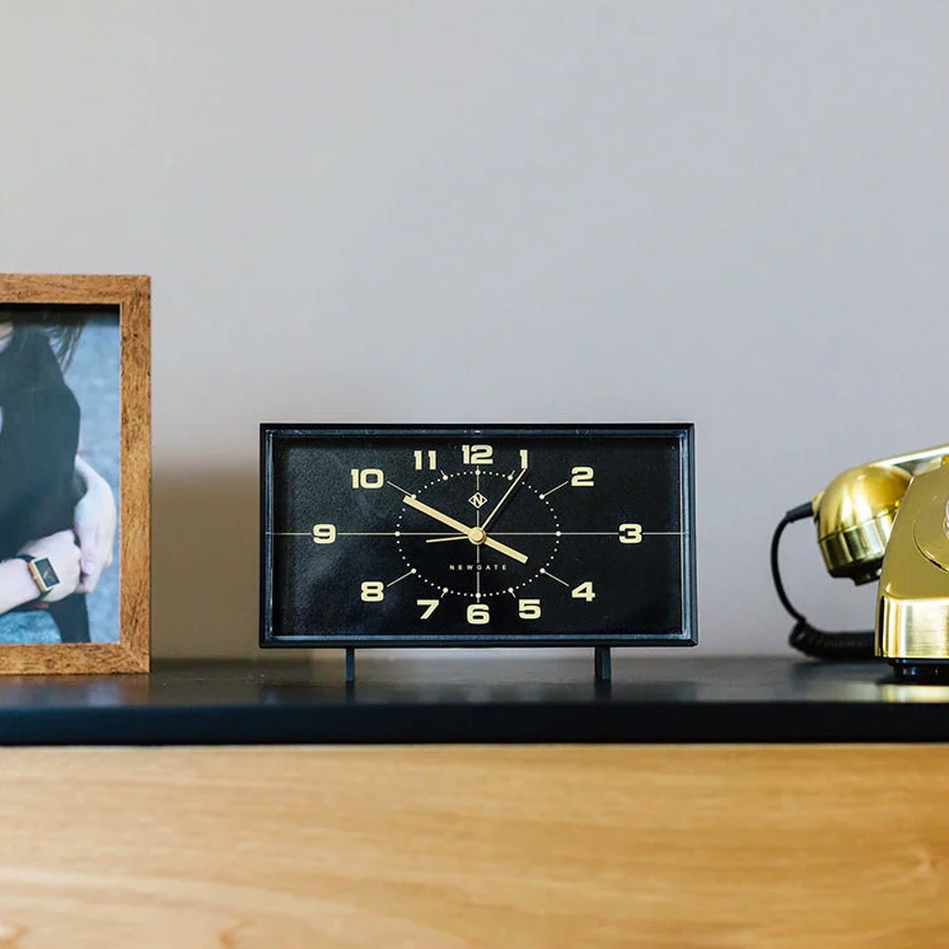 The Newgate Wideboy Alarm Clock by Newgate, featuring a mid-century atomic dial and gold numbers, is displayed on a wooden surface beside a framed photo and a vintage gold rotary phone.