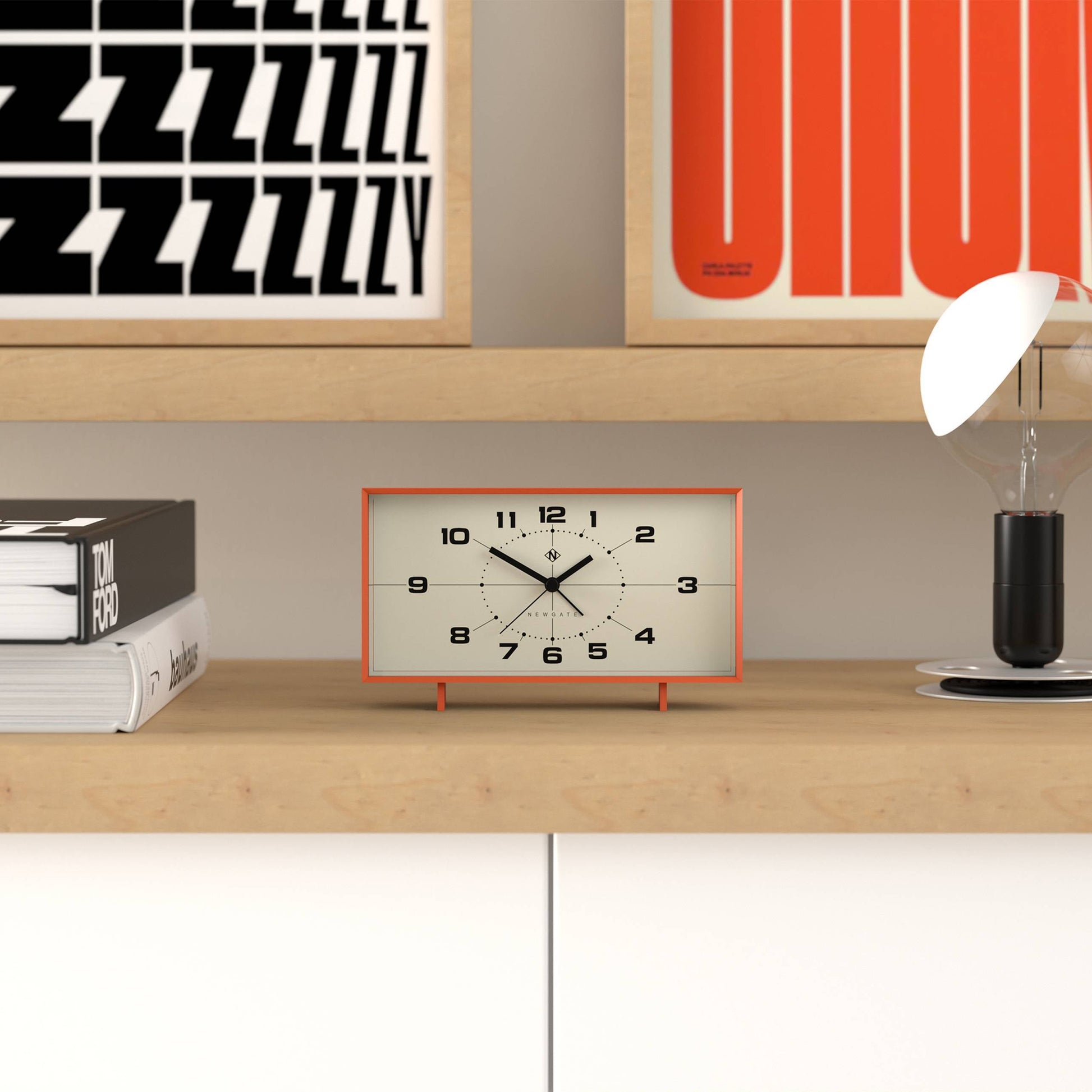 A rectangular orange and white Newgate Wideboy Alarm Clock by Newgate sits on a wooden shelf beside a light bulb lamp, books, and colorful graphic artwork displayed on the wall in the background.