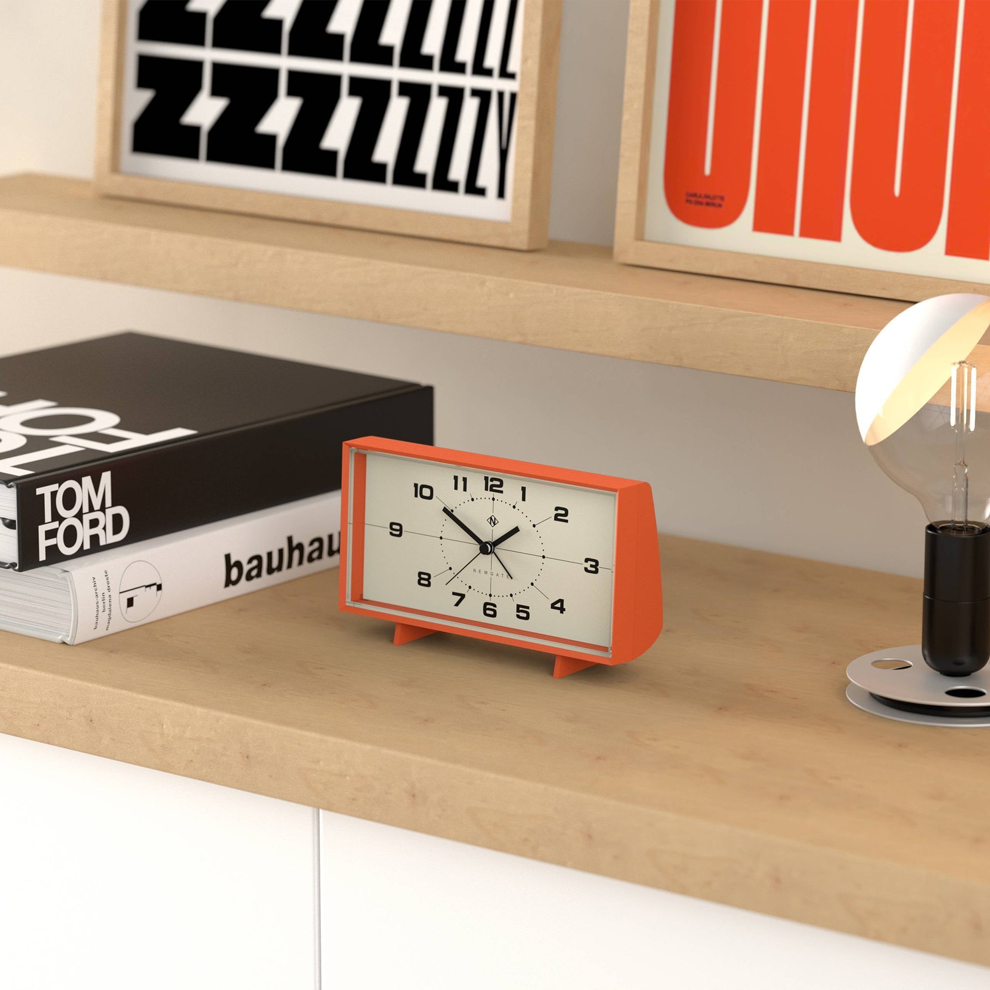 A Newgate Wideboy Alarm Clock with a mid-century atomic dial sits on a wooden shelf alongside design books—including one titled Tom Ford—a framed graphic print, and a modern lightbulb.